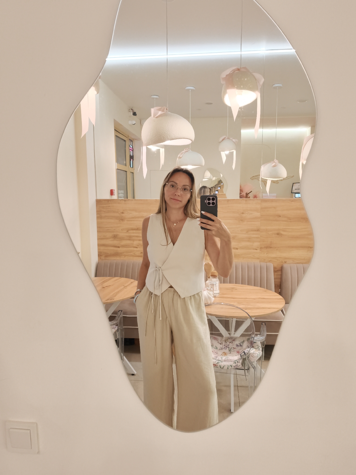 A woman with blonde hair and glasses, wearing a white vest and beige wide-leg pants, stands in a cafe and takes a mirror selfie. The cafe has light-colored walls, wooden accents, and round tables with transparent chairs that have floral patterned cushions. Unique light fixtures with pink ribbon accents hang from the ceiling. The mirror itself has an irregular, wavy shape. The scene appears to be captured in a modern, stylish cafe.