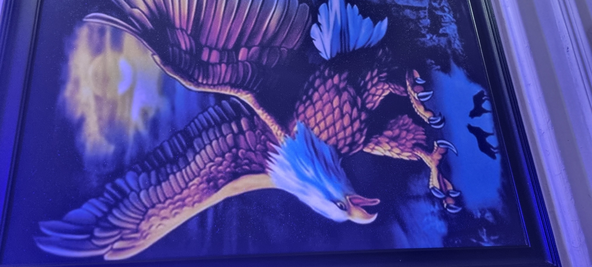 A framed painting hangs on a wall, depicting a large, stylized eagle in flight. The eagle is rendered in shades of purple, blue, and orange. The head is blue, with a white beak. The body appears to be covered in scale like plumage. The wings are spread wide, with feathers detailed in shades of purple and gold. In the background, there is a hint of light, possibly from a moon or sun, and the shadows of other birds flying. The painting is framed in a black frame. The location is unknown, and Aiyetoto-Asogun, Nigeria cannot be confirmed.
