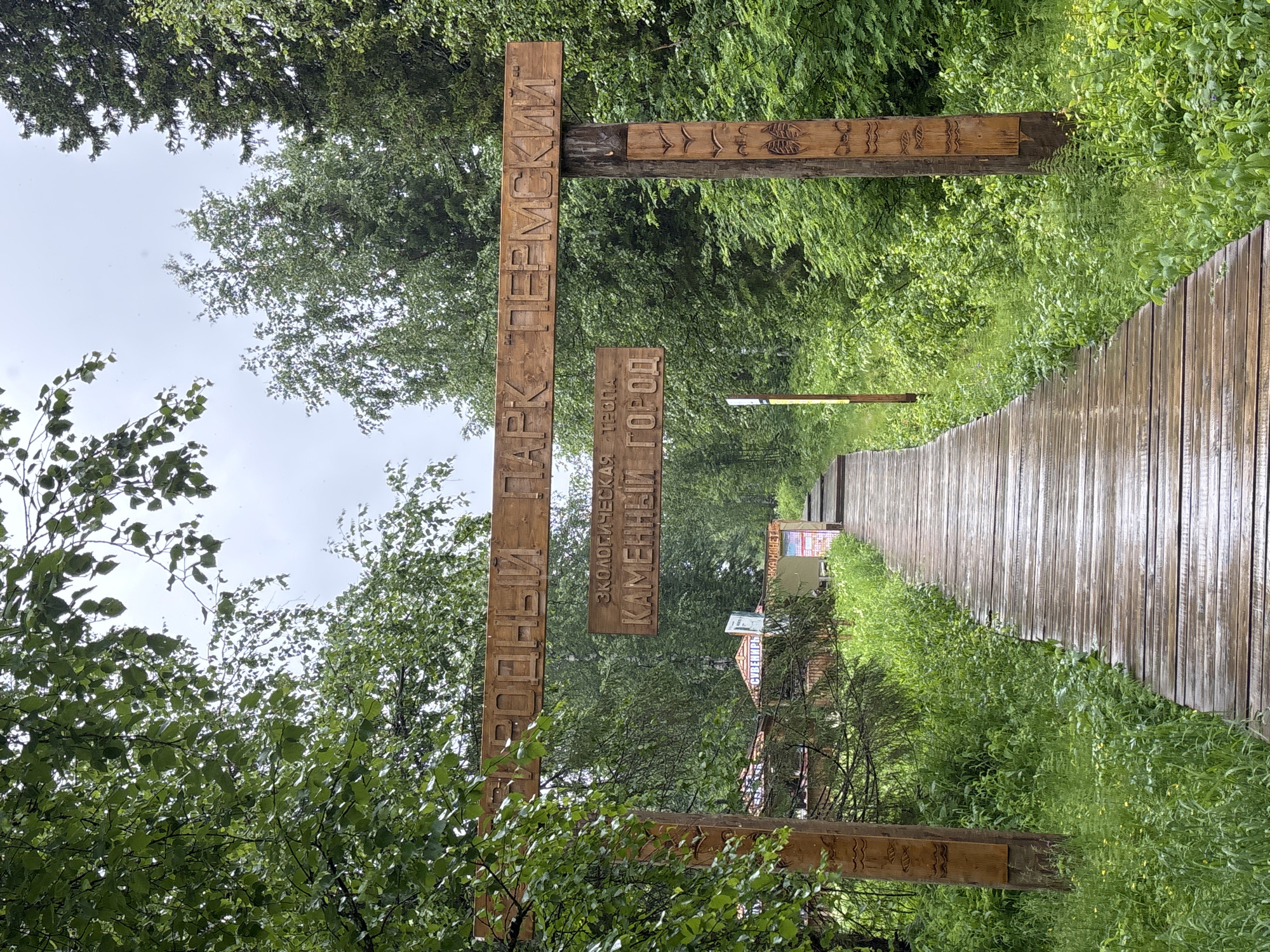 The image shows a wooden archway with carved inscriptions at the entrance to a park. The archway is made of rough-hewn wood and is decorated with carvings of birds and fish. The inscriptions on the archway read "ПРИРОДНЫЙ ПАРК ПЕРМСКИЙ" and "ЭКОЛОГИЧЕСКАЯ ТРОПА КАМЕННЫЙ ГОРОД". Beyond the archway, a wooden boardwalk leads into a lush, green forest. The boardwalk is wet, suggesting it may have recently rained. Trees and dense foliage line both sides of the path. In the background, there are small wooden structures that appear to be part of the park's facilities. The overall scene depicts a natural park entrance, likely in the region of Usva, Russia, given the inscription "ПРИРОДНЫЙ ПАРК ПЕРМСКИЙ". There are no people visible in the image.