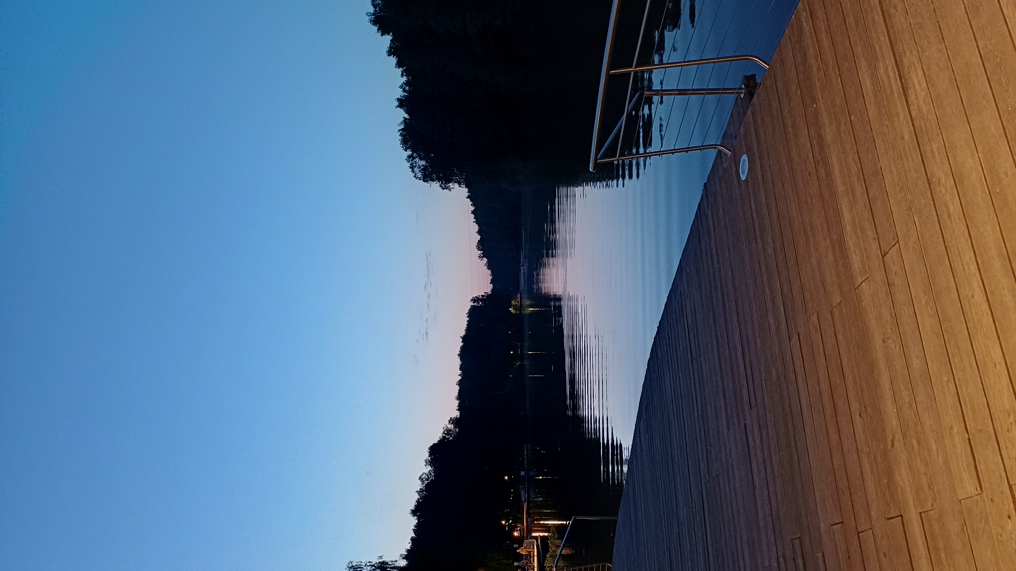 The image depicts an evening scene in Balashikha, Russia, featuring a wooden deck alongside a body of water, likely a lake or river. The deck extends towards the water, with a stainless-steel railing visible along its edge. The water reflects the twilight sky, which transitions from a light blue to a soft pink hue towards the horizon. Trees line the opposite bank, appearing dark against the illuminated sky. Lights are visible along the shoreline, suggesting a populated area. The wooden deck is illuminated by warm lighting, contrasting with the cool tones of the sky and water.