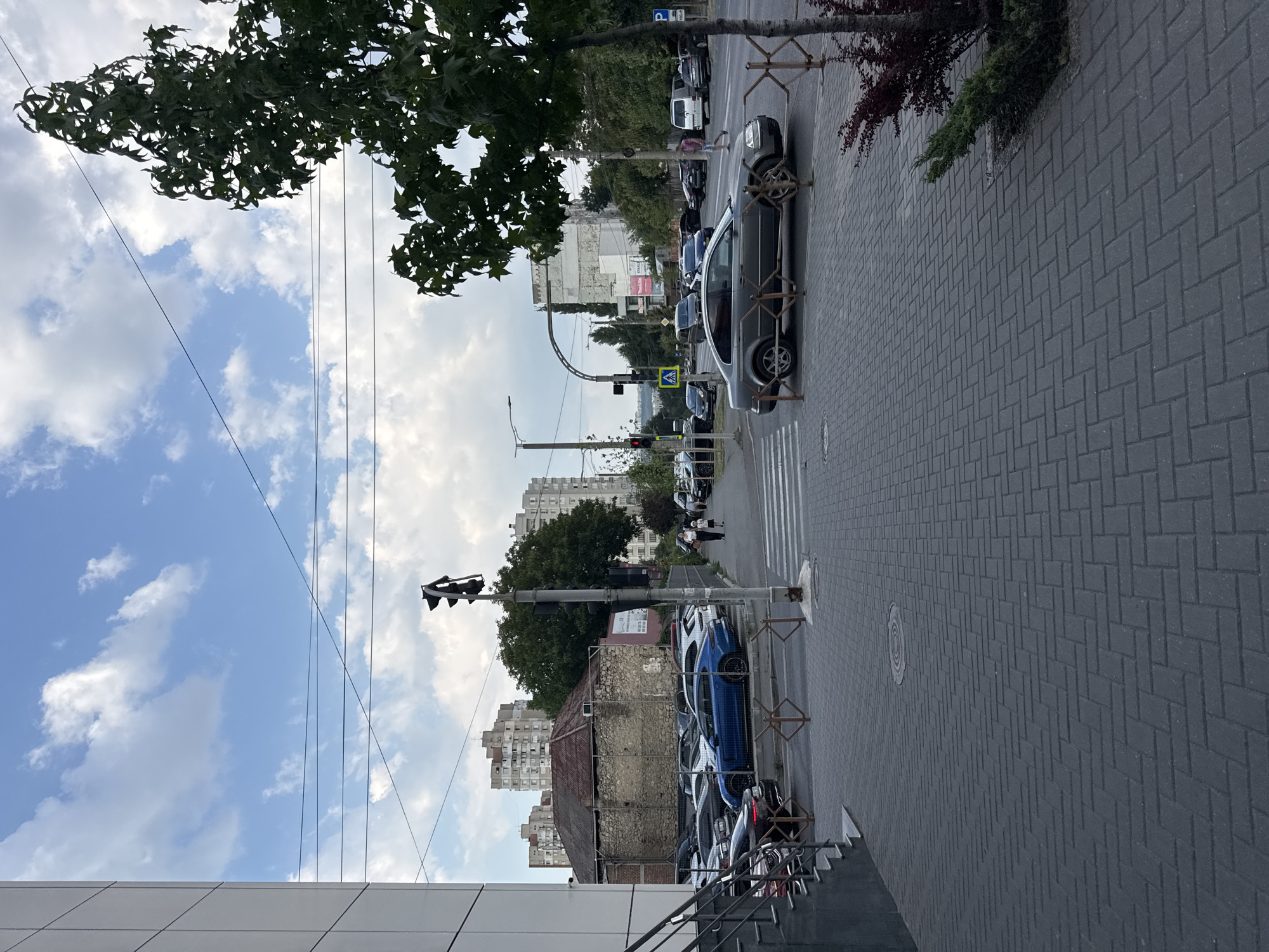 This image depicts a street scene in Chișinău, Moldova. On the left, a modern building with a white facade and visible metal framing dominates the foreground. A gray staircase leads up to its entrance. Adjacent to the modern building, an older, stone-built structure with a red-tiled roof stands out. A parking lot filled with numerous cars, including a prominent blue sedan, is situated behind a low metal fence.
The street itself is paved with dark gray rectangular cobblestones. A pedestrian crossing is clearly marked, with metal barriers delineating its edges. Several cars are visible on the street, including a dark gray sedan in the foreground. Traffic lights are present, with one showing a red light. A few pedestrians are walking on the sidewalk, one a woman with a child.
The background features a cluster of multi-story apartment buildings, typical of Soviet-era architecture. Trees are scattered throughout the scene, with a large, leafy tree on the right side of the image. The sky is a mix of blue and white clouds. Power lines are strung across the sky. The overall impression is that of an urban environment in daylight.