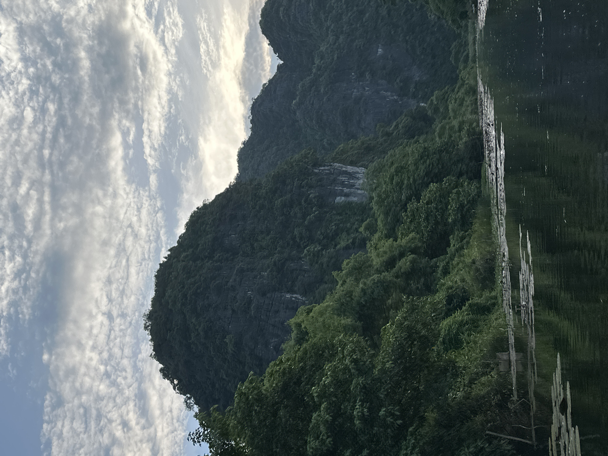 A body of water reflects the lush green mountains that surround it. Lily pads float on the surface of the water, and trees grow on the slopes of the mountains. The sky is partly cloudy, with the sun peeking through the clouds. The scene appears to be in Nam Hoa Lư Ward, Vietnam, a region known for its karst landscape and tranquil waterways. There are no people or animals visible in the image.