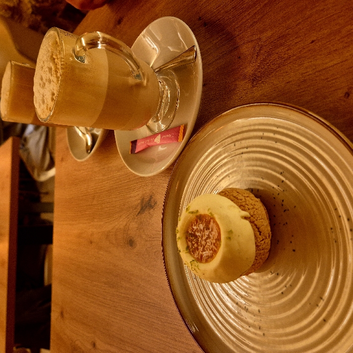 A tan wooden table features a dessert on a ribbed, round plate with a patterned edge. The dessert has a light yellow outer layer and a textured center. A glass mug of light brown frothy liquid sits on a saucer with a spoon. Another similar beverage is partially visible in the background. A pink packet lies on the saucer. The composition is warm-toned.