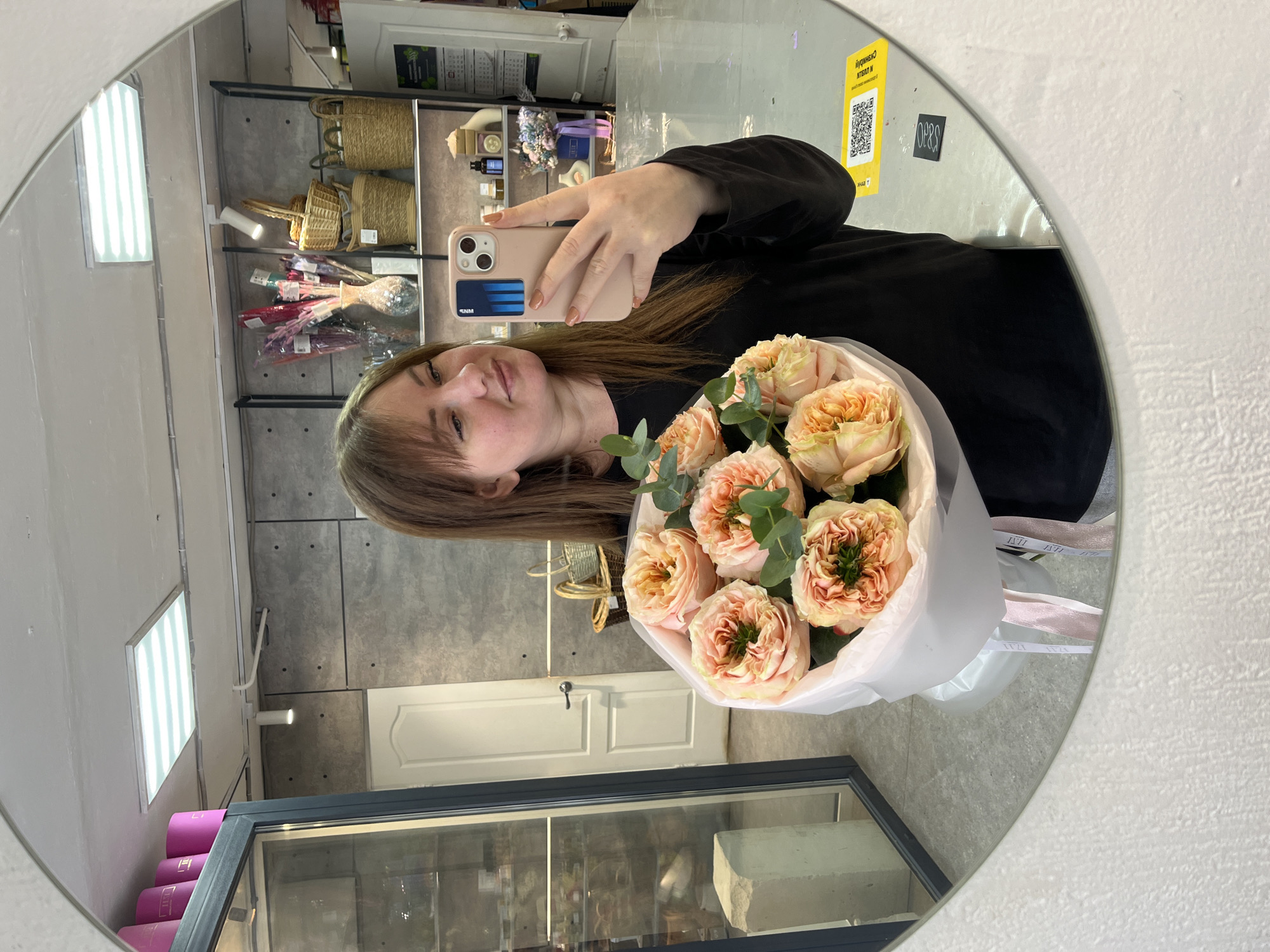 A selfie in a round mirror shows a woman with shoulder-length brown hair, bangs, and fair skin. She is wearing a black long-sleeved shirt and holding a bouquet of light orange roses with green leaves. She holds up a tan-colored smartphone in her left hand. The background includes shelves with baskets and other items, a glass display case with products, and a white door in the gray wall. A yellow sticker with QR code is seen attached to the mirror. Based on the "IZI" inscription on the bouquet ribbons and QR code on the yellow sticker, the photo likely was taken at the flower shop "IZI FLOWER" in the city of Chaikovsky, Russia.