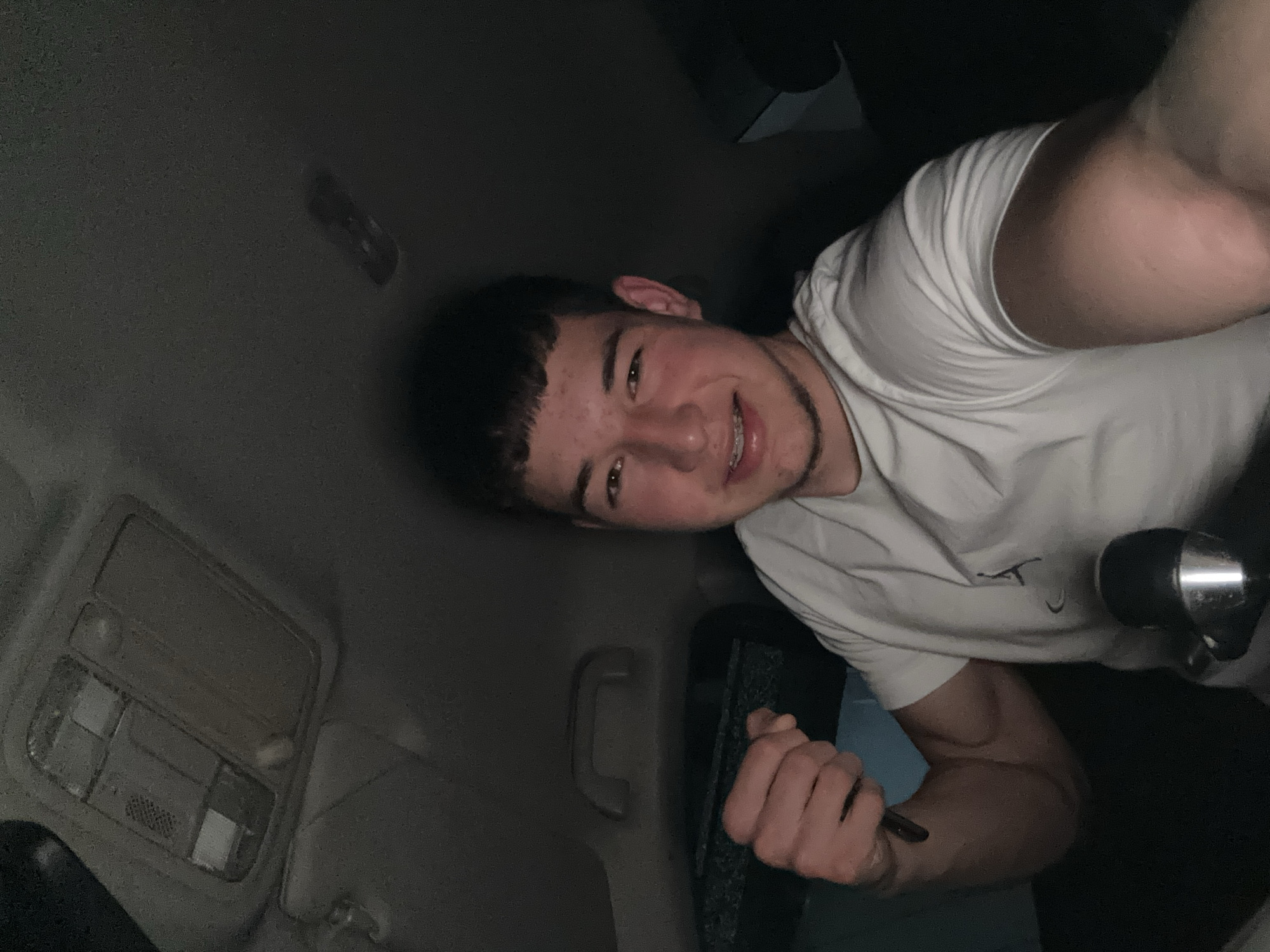 A young man, likely in his late teens or early twenties, is captured in a close-up, informal portrait taken inside a car or SUV, most probably at night or in a dimly lit environment. He is looking directly at the camera with a wide, friendly smile, showcasing braces on his upper teeth. He has short, dark hair and appears to have a few freckles or minor blemishes on his forehead.
He is wearing a plain white short-sleeved t-shirt, which features a small, indistinct dark graphic or logo on the chest area. His right arm is visible, showing a muscular build, and his right hand is holding a thin, dark, pen-like object. The car's interior is identifiable by the light-colored headliner, an overhead console with lights and controls on the left, and a grab handle above what appears to be a window. Another dark object, possibly part of a car seat or interior trim, is visible near his right shoulder. The overall mood is casual and cheerful. No legible text is visible within the image itself.