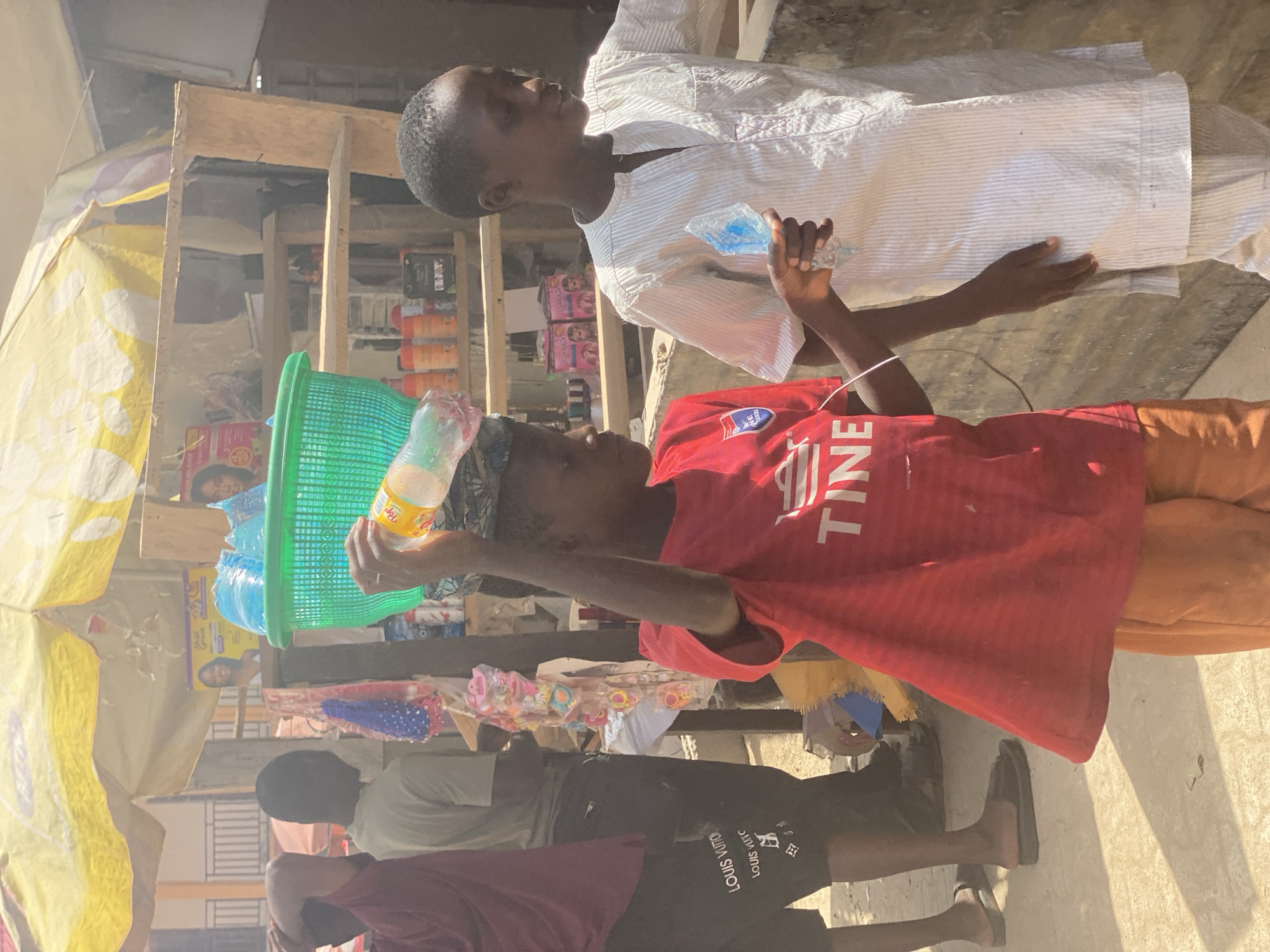 A bustling street scene in Kafanchan B, Nigeria, features several individuals and various objects. A young person wearing a red t-shirt with the word "TINE" visible is positioned near a green plastic basket filled with bottled water. Another person in a white striped shirt stands nearby, holding a plastic water bottle. The scene takes place near a market stall or shop with various goods displayed on shelves. Two more people are standing in the shade in front of the stall. The weather appears sunny, and the overall atmosphere suggests a typical day in a commercial area.