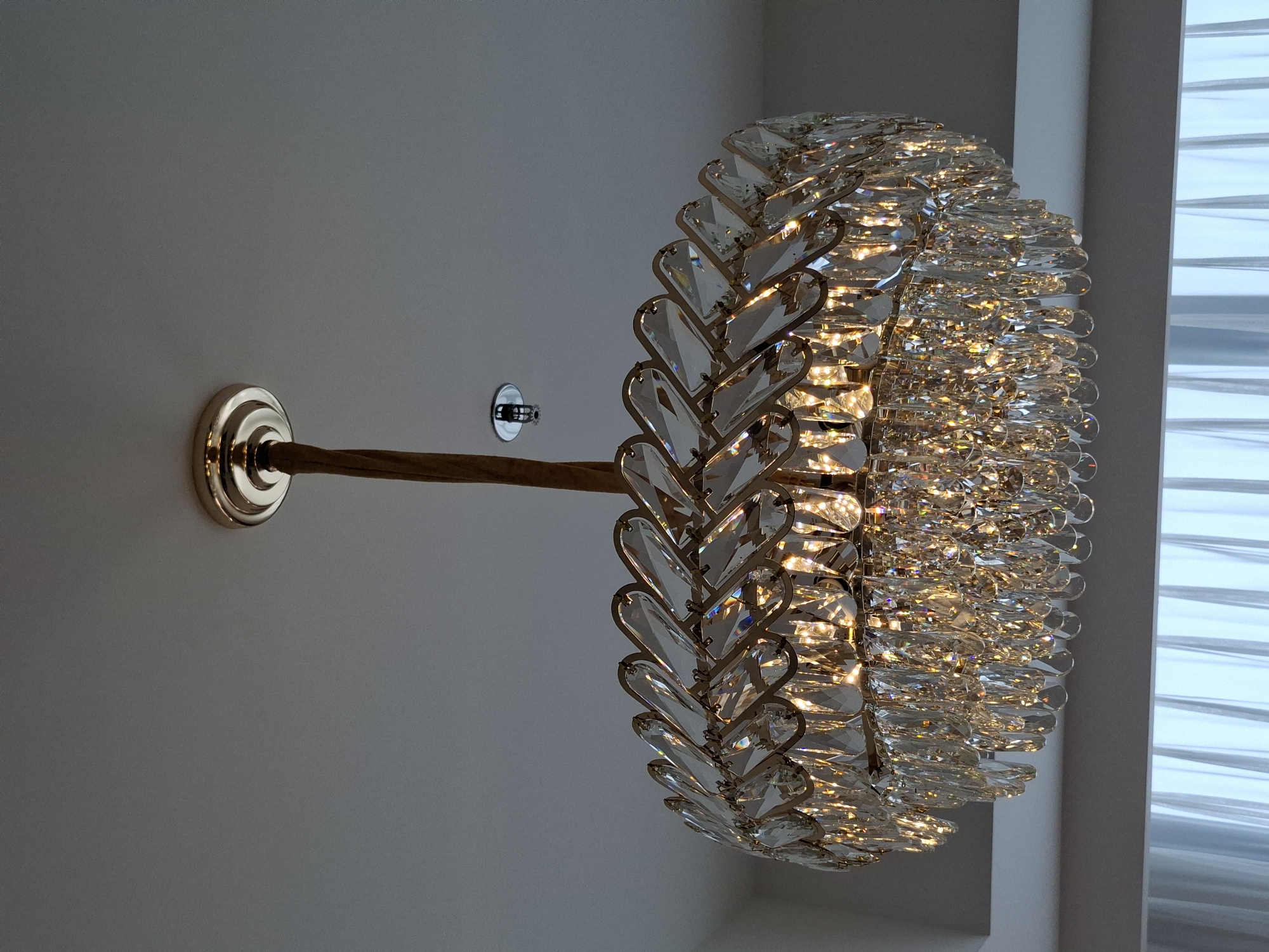 A crystal chandelier hangs from a white ceiling, suspended by a braided brown cord. The chandelier is round and features multiple tiers of clear, faceted crystals that reflect light. The crystals are teardrop-shaped and arranged in a layered, opulent design. Above and to the left of the chandelier, a silver fire sprinkler head is visible mounted on the ceiling. In the background, a light-colored curtain is partially visible, suggesting the presence of a window. The image appears to be taken indoors. The location in the city of West Jakarta, Indonesia, is not explicitly depicted in the visual information provided.