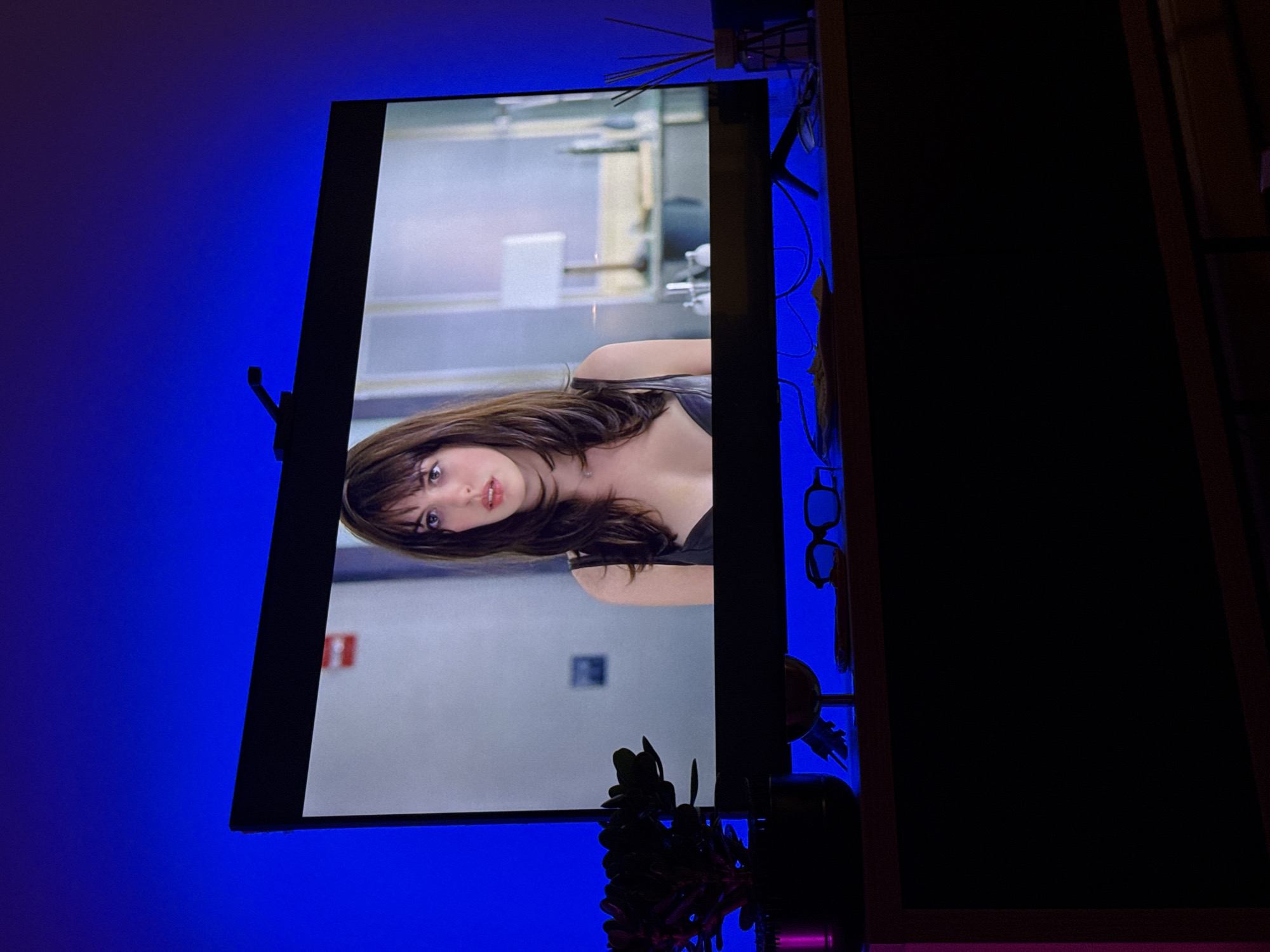 A flat-screen television displays a medium shot of Anne Hathaway, apparently from the movie "Love & Other Drugs," showing her in a lab coat. The TV is mounted on a shelf or stand. The background behind and around the TV is illuminated with blue LED lights, creating a bright blue backdrop. The right side of the image is dominated by a dark, rectangular object, likely part of the shelf or stand the TV is on. A plant is also visible at the bottom of the screen. There is no information about the location in Belgrade, Serbia within the visible image.