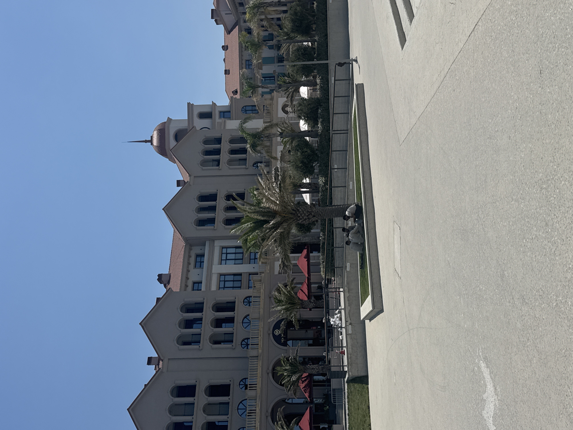A wide shot captures a multi-story building with a Mediterranean architectural style, featuring arched windows and red tile roofs, in Sabunchu Raion, Azerbaijan. The building appears to be a hotel or resort, with palm trees and red umbrellas in front of it. In the foreground, a group of four men are sitting on a patch of grass, appearing to converse. The ground is paved with light-colored concrete, and there is a clear blue sky overhead.