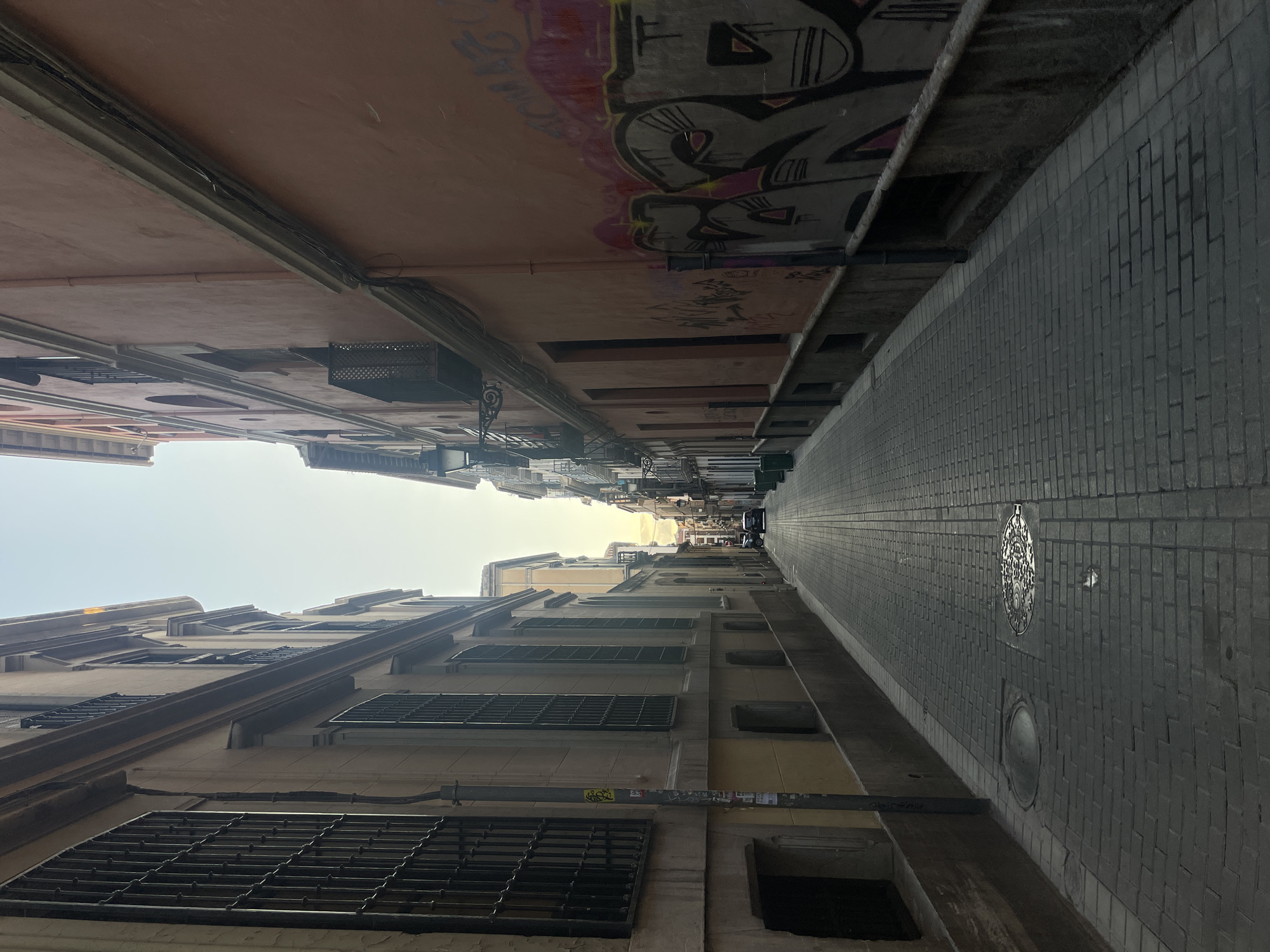 The image depicts a narrow street in Granada, Spain, likely within the Albaicín district, characterized by its historical architecture and cobblestone pavement. On the left, a building with beige-colored walls features tall windows protected by black wrought-iron bars. A gray metal pole with some stickers is prominent near the foreground. The building on the right is painted in a salmon or pink hue and is adorned with graffiti in various colors, including pink, gray, and black. This side of the street also has several windows and balconies with black railings. The street itself is paved with uniform gray bricks, and two manhole covers are visible in the foreground, one round and one square with an intricate pattern. A drainpipe runs vertically along the pink wall. In the distance, the street curves slightly and narrows, with more buildings visible, some with lampposts and balconies. A car is parked further down the street on the right side, and another vehicle is in motion, heading away from the camera. The sky above is a pale, hazy blue, suggesting it is either early morning or late afternoon, with sunlight casting a soft glow. The overall impression is of an old European city street, with a blend of historical charm and urban elements.