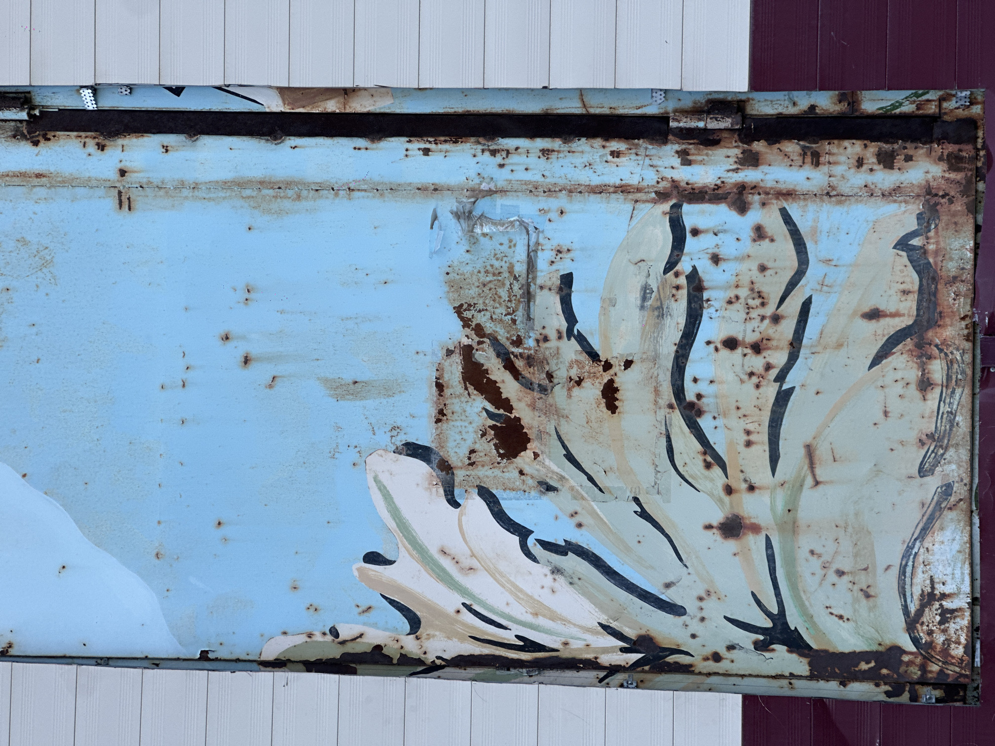 An aged, rusted metal panel sits adjacent to a wall composed of both white and maroon siding. The panel is light blue with an off-white stylized floral pattern. Rust is present across the panel, obscuring portions of the painted design. Sayansk, Russia, cannot be confirmed.