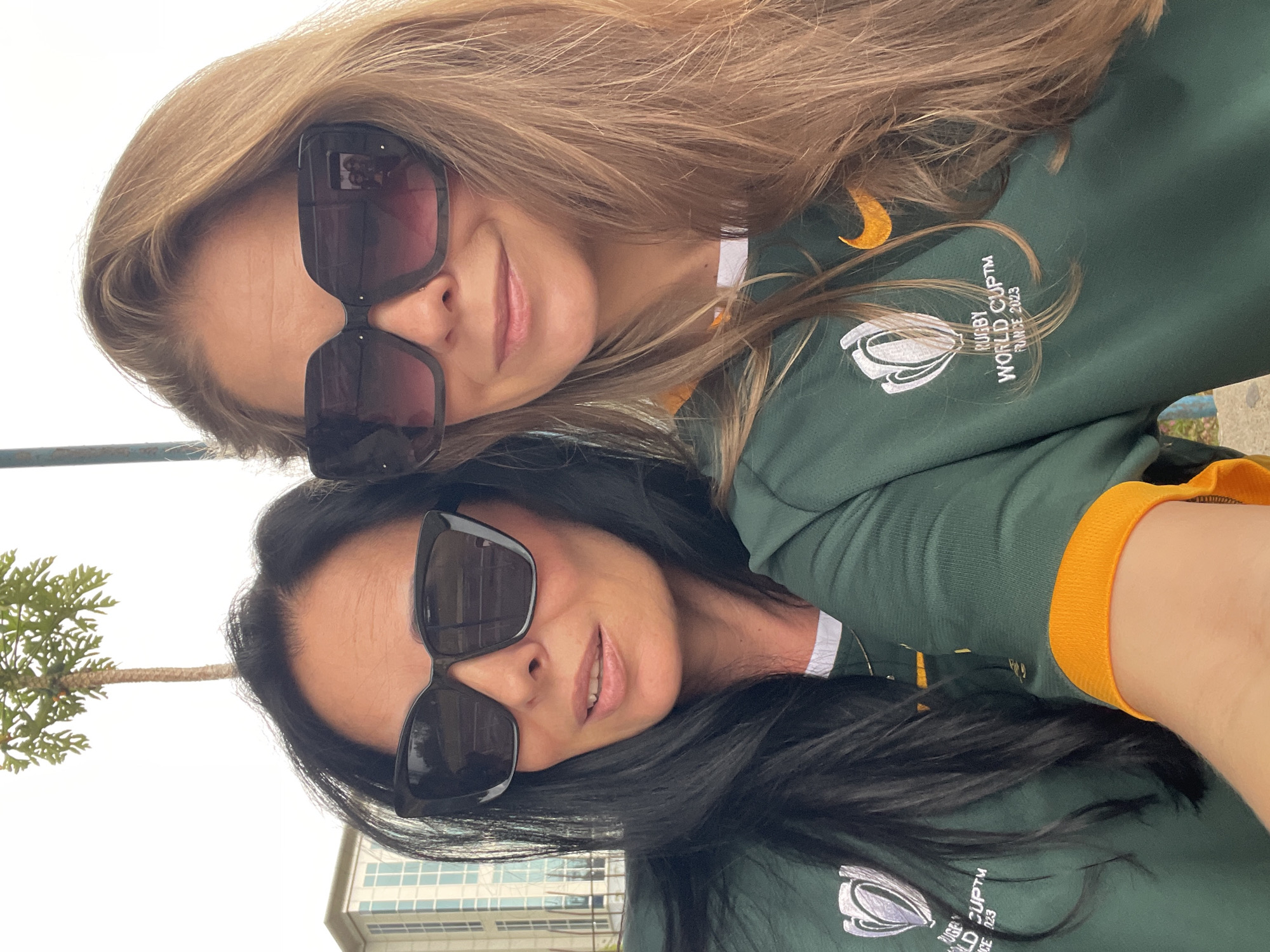 The image shows two women in sunglasses, wearing green rugby jerseys. The woman on the left has dark hair and the woman on the right has blonde hair. They are both smiling and looking at the camera. The jerseys have the Rugby World Cup France 2023 logo on them. In the background, there is a building with blue windows and a palm tree. The scene appears to be outdoors, possibly in a park or public space. The lighting suggests it is daytime. The location is not discernible from the image itself.