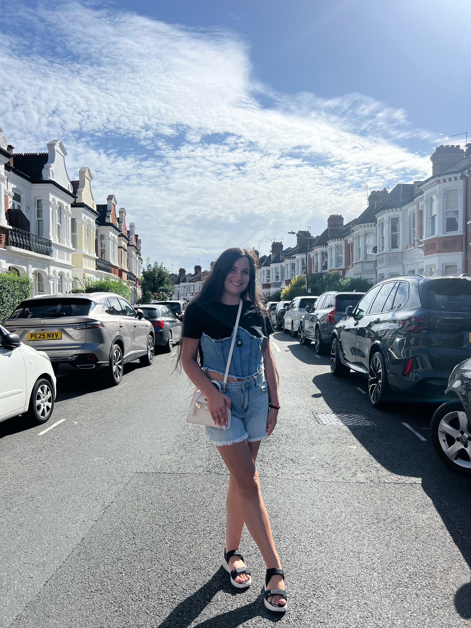 A young woman with long dark hair and a smiling expression stands on a residential street in London. She is wearing denim overalls over a black t-shirt and black and white sandals. She is carrying a small white purse. The street is lined with parked cars and Victorian-style houses. The sky is blue with scattered white clouds. The sun is shining brightly, casting shadows on the street. The license plate of the car in the foreground reads "PE25 NVV".