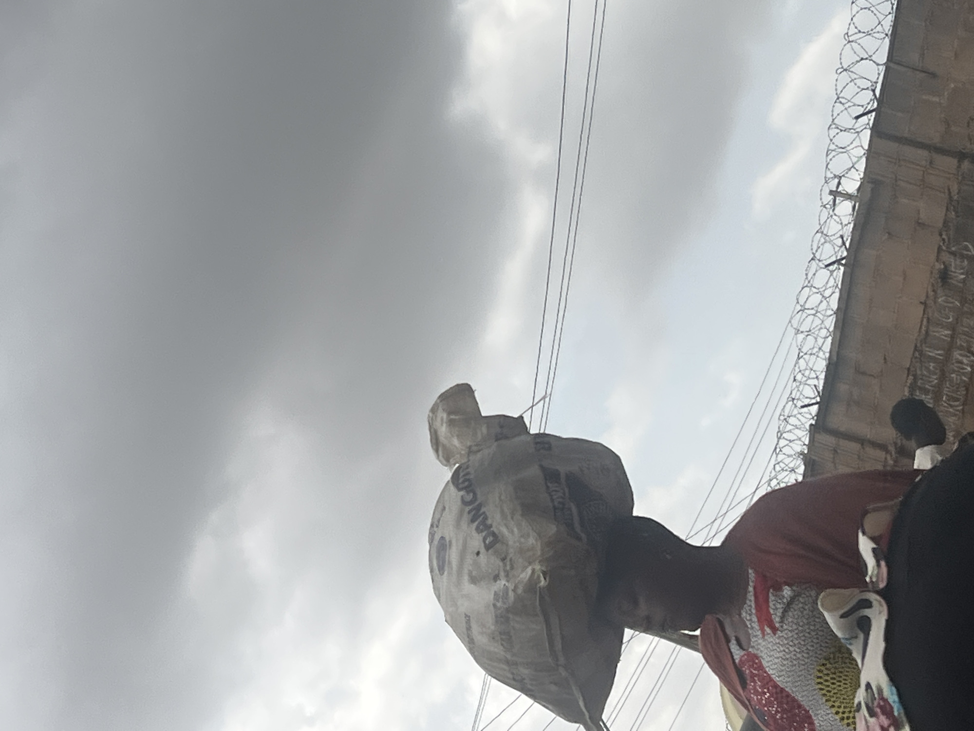 A person is carrying a large bag of "Dangote" cement on their head. The sky is overcast. A wall with barbed wire on top is visible in the background. The location is suggested to be Gura Top, Nigeria.