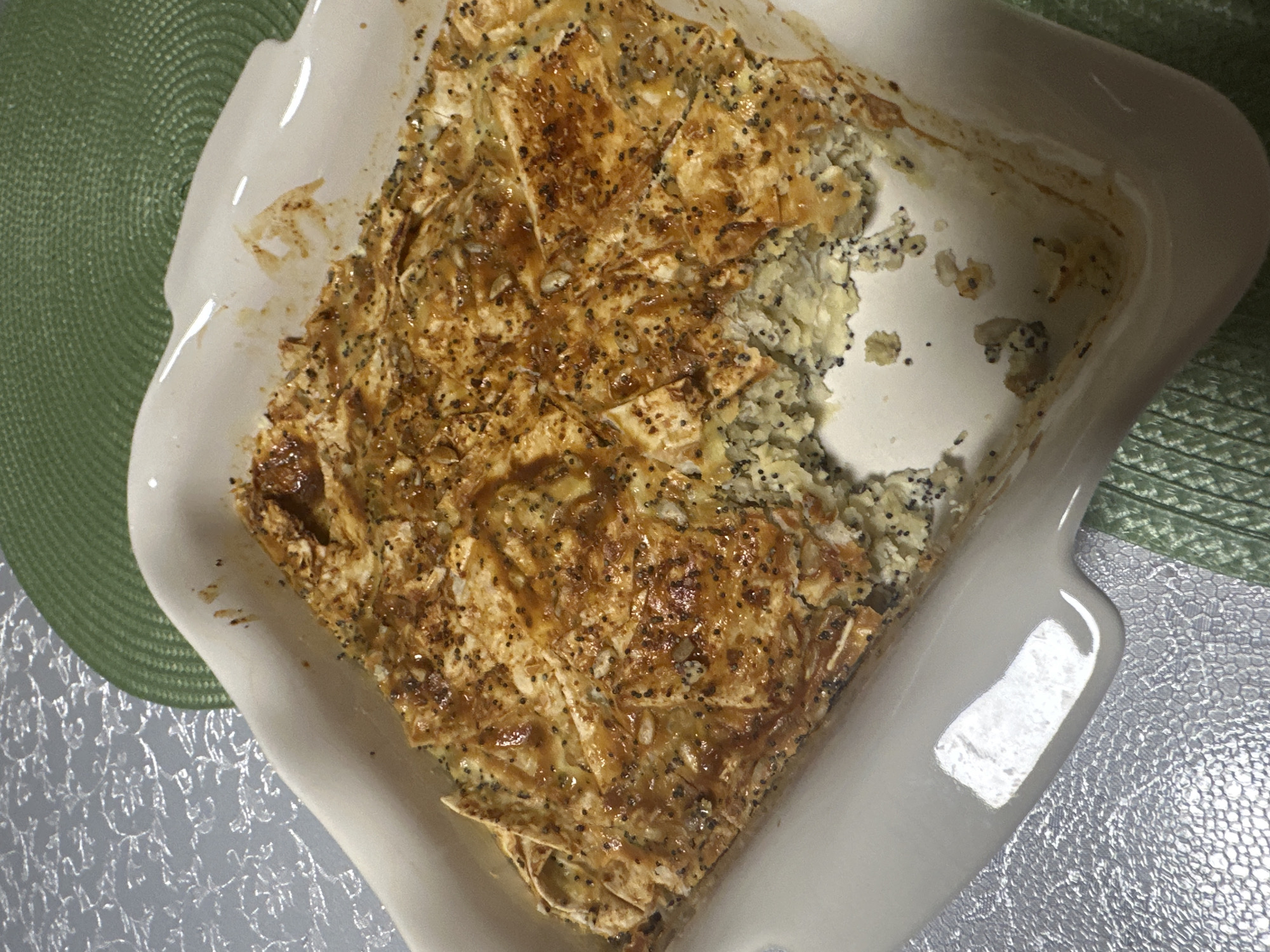 A partially eaten poppy seed pastry is in a white, square baking dish with rounded corners. The dish is sitting on a silver, textured surface. A green, round woven placemat is partially visible on the left side of the dish, and a green placemat is on the right side. The pastry has a golden-brown, flaky crust with poppy seeds scattered on top. The filling appears to be a light-colored mixture with poppy seeds. A portion of the pastry is missing from the top right. The baking dish is slightly stained on the interior. The location is not discernible in the image.
