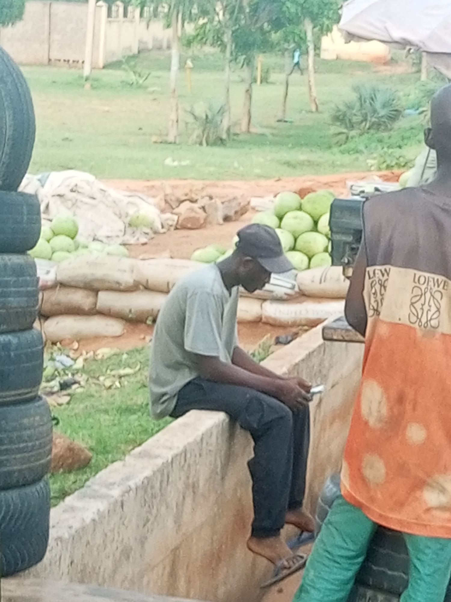 The image depicts an outdoor scene in Garko, Nigeria, likely a marketplace or roadside stall. In the foreground, two individuals are partially visible. One person, wearing a grey t-shirt and dark pants, is seated on a low concrete wall, looking down at a mobile phone in their hands. They are wearing a dark baseball cap and sandals. To the right, another person, seen from behind, is wearing an orange and grey shirt with visible "LOEWE" branding and green pants.
Behind the seated individual, a stack of dark tires is visible on the left side of the frame. Further back, a large pile of light green, melon-like fruits, possibly watermelons or cantaloupes, are arranged. These fruits are placed on top of several large, beige sacks, which appear to be bags of cement or other bulk goods, stacked on the ground. The ground surface is a mix of dirt and sparse green grass. In the background, there are trees and some fencing, suggesting a semi-rural or peri-urban environment. The overall lighting suggests it is daytime, possibly late morning or afternoon. No specific weather conditions are evident. The scene appears to be one of commerce, with goods being displayed and sold.