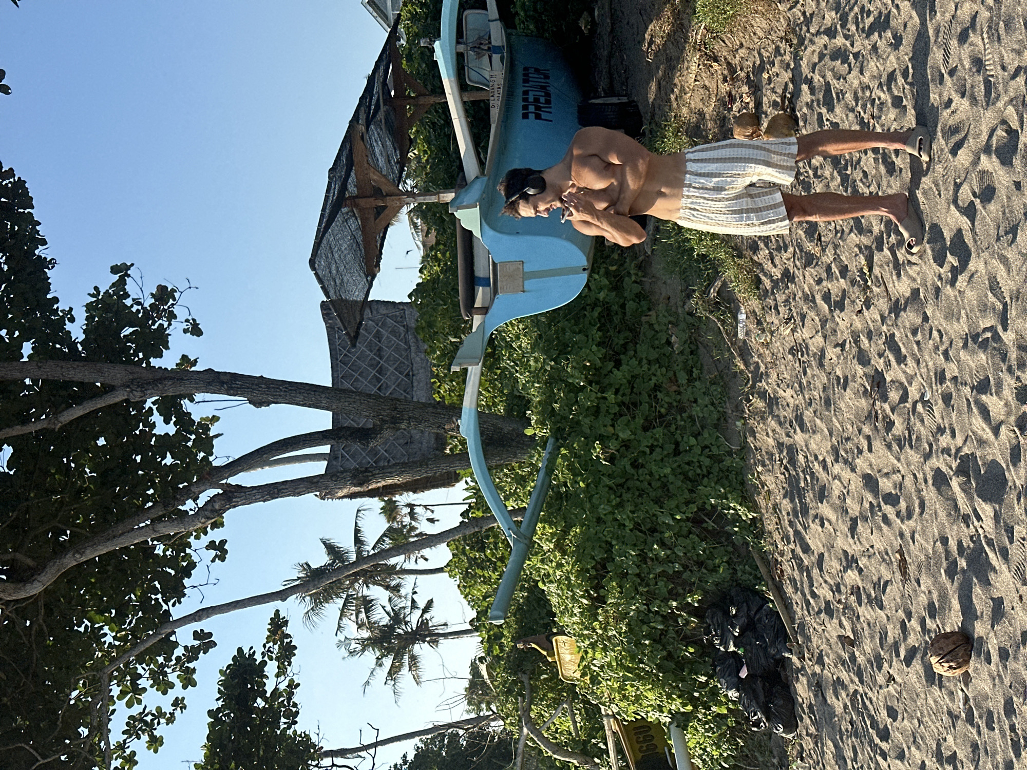 A shirtless man with headphones stands on a sandy beach in North Kuta, Indonesia, next to a blue boat with the word "PREDATOR" on its side. The man is wearing patterned shorts and appears to be holding something near his mouth. Behind him, lush green foliage and palm trees are visible. In the foreground, a coconut sits on the sand. Several black trash bags are also piled on the sand to the left of the man. The sky is clear and blue.