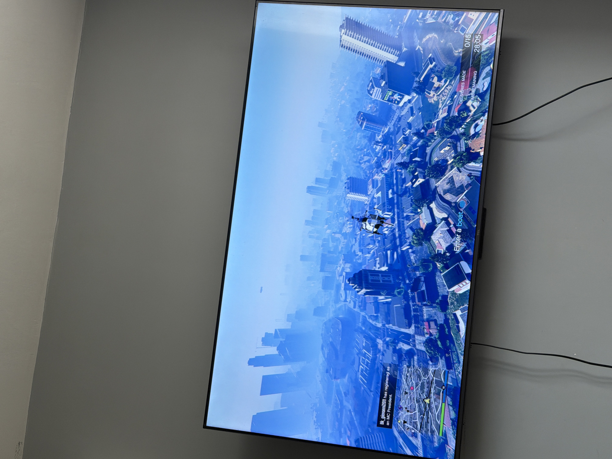 A television screen, mounted vertically on a light gray wall, displays a video game scene. The game appears to be an open-world title, likely Grand Theft Auto V, viewed from an aerial perspective over a sprawling, hazy cityscape. Skyscrapers and urban infrastructure are visible below a clear, bright blue sky with a slight atmospheric haze. A character, possibly a drone or a person skydiving, is central to the view.
On the game's interface, various elements are visible. In the upper right corner, an objective tracker shows "DELIVERIES MADE 0/15" and "TIME REMAINING 28:05", indicating a mission in progress. At the bottom of the screen, a minimap with navigation points and other in-game icons is displayed. Above the minimap, a notification reads "Bk_giovanni2011 has registered as an MC President." Another prompt near the bottom asks the player to "Enter a boat". The time of day within the game is clearly daytime, characterized by bright lighting.
To the right of the TV, a dark power cable runs horizontally across the wall. The wall itself is painted a uniform, light gray color. The image is taken from an angle, causing the television to appear rotated by 90 degrees clockwise relative to how it would normally be viewed.