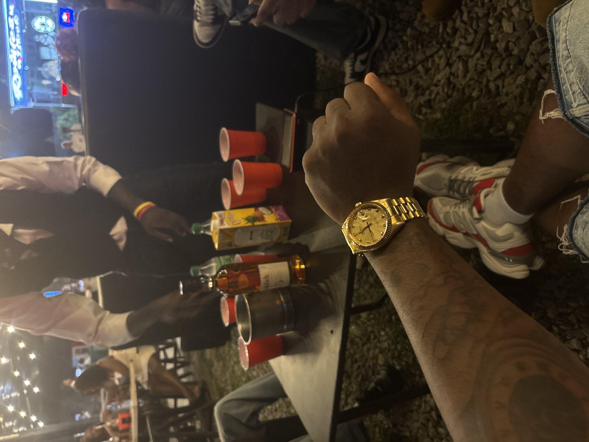The image captures an outdoor nighttime social gathering, likely a party, set in Abuja, Nigeria. The perspective is from a person's point of view, prominently featuring their left forearm adorned with intricate tattoos and a gold watch, possibly a Rolex Day-Date, with the text "ROLEX OYSTER PERPETUAL DAY-DATE SUPERLATIVE CHRONOMETER OFFICIALLY CERTIFIED" and "SWISS MADE" visible on its dial.
In the foreground and midground, a dark table is central to the activity. It holds several red plastic cups, some stacked or empty, alongside bottles of spirits, including one identifiable as "BELL'S" (likely Bell's Original Blended Scotch Whisky), and a "PINEAPPLE" juice box (resembling Cappy or Capri-Sun). Another person's legs are visible to the right, wearing denim shorts with rips and white and red sneakers.
In the background, other individuals are seen sitting or standing around the table, engaged in conversation or drinking. The setting is illuminated by decorative string lights and a bright, light-up sign in the far background, indicating the nighttime environment. The ground appears to be gravel or crushed stone, typical of an outdoor venue. The overall atmosphere suggests a casual and relaxed social event.