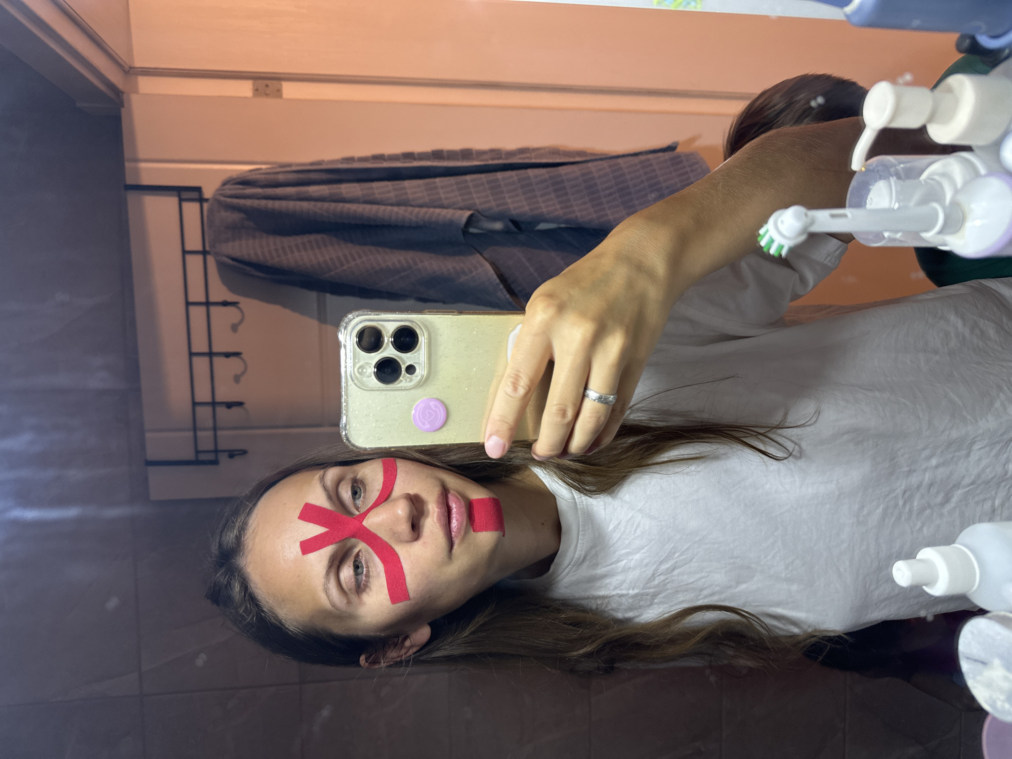 A woman is taking a selfie in a bathroom. She has red kinesiology tape on her face in an "X" shape across her forehead and a small piece on her upper lip. She is wearing a white t-shirt and has long, brown hair. She is holding a smartphone with a clear case. In the background, there is a towel rack with a gray towel, various bathroom toiletries, and a mirror. The location is not specified and cannot be determined from the image.