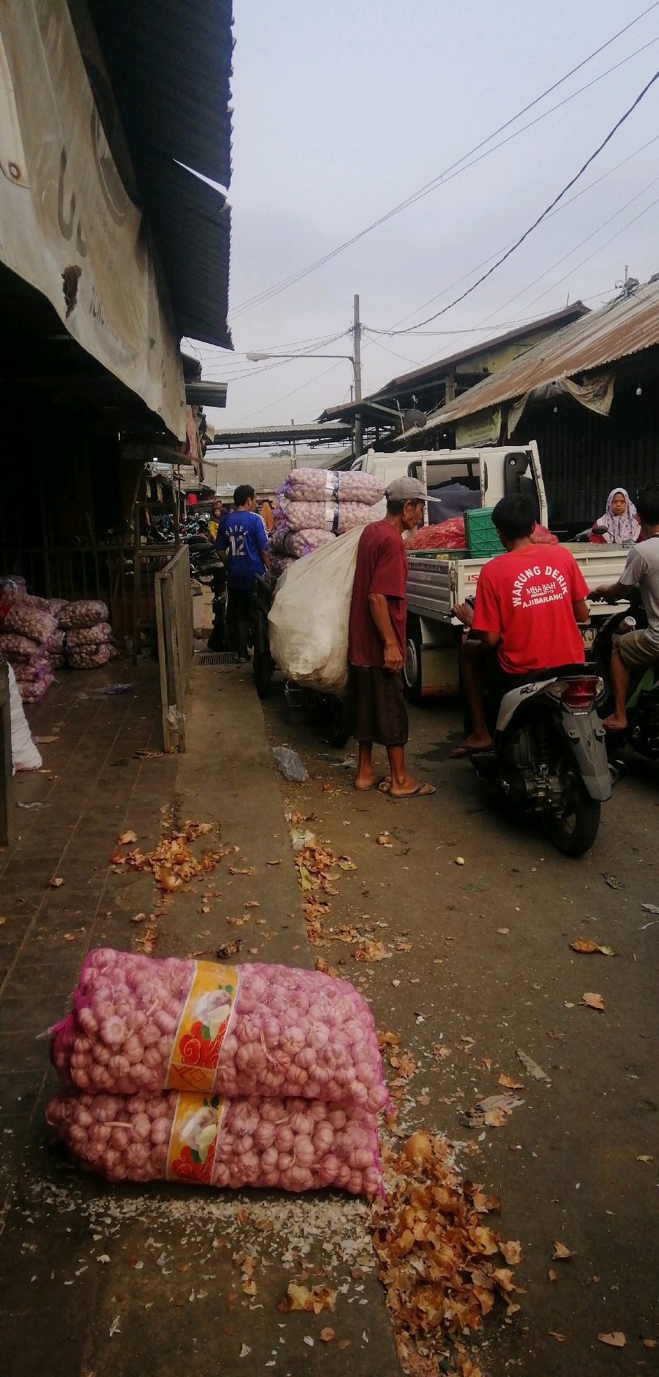 The image depicts an outdoor market scene in Ajibarang, Indonesia. The foreground is dominated by two large, pink mesh bags filled with garlic, resting on the ground. Scattered around the garlic bags are dried husks and debris, suggesting a busy marketplace.
In the mid-ground, a white truck is parked, its bed loaded with numerous large, tightly packed sacks of produce, likely also garlic, wrapped in white plastic. Several people are present. A man in a red t-shirt with the text "WARUNG DERIC MBAH AJIBARANG" on the back is sitting on a scooter. To his left, another man in a maroon t-shirt and dark shorts is standing, carrying a large, light-colored sack over his shoulder. Further back, a man in a blue t-shirt with the number "12" is standing near a motorcycle.
In the background, there are market stalls with corrugated metal roofs, some appearing weathered and dilapidated. Power lines and a street lamp are visible against an overcast sky. The overall impression is a bustling, possibly somewhat disorganized, agricultural market.