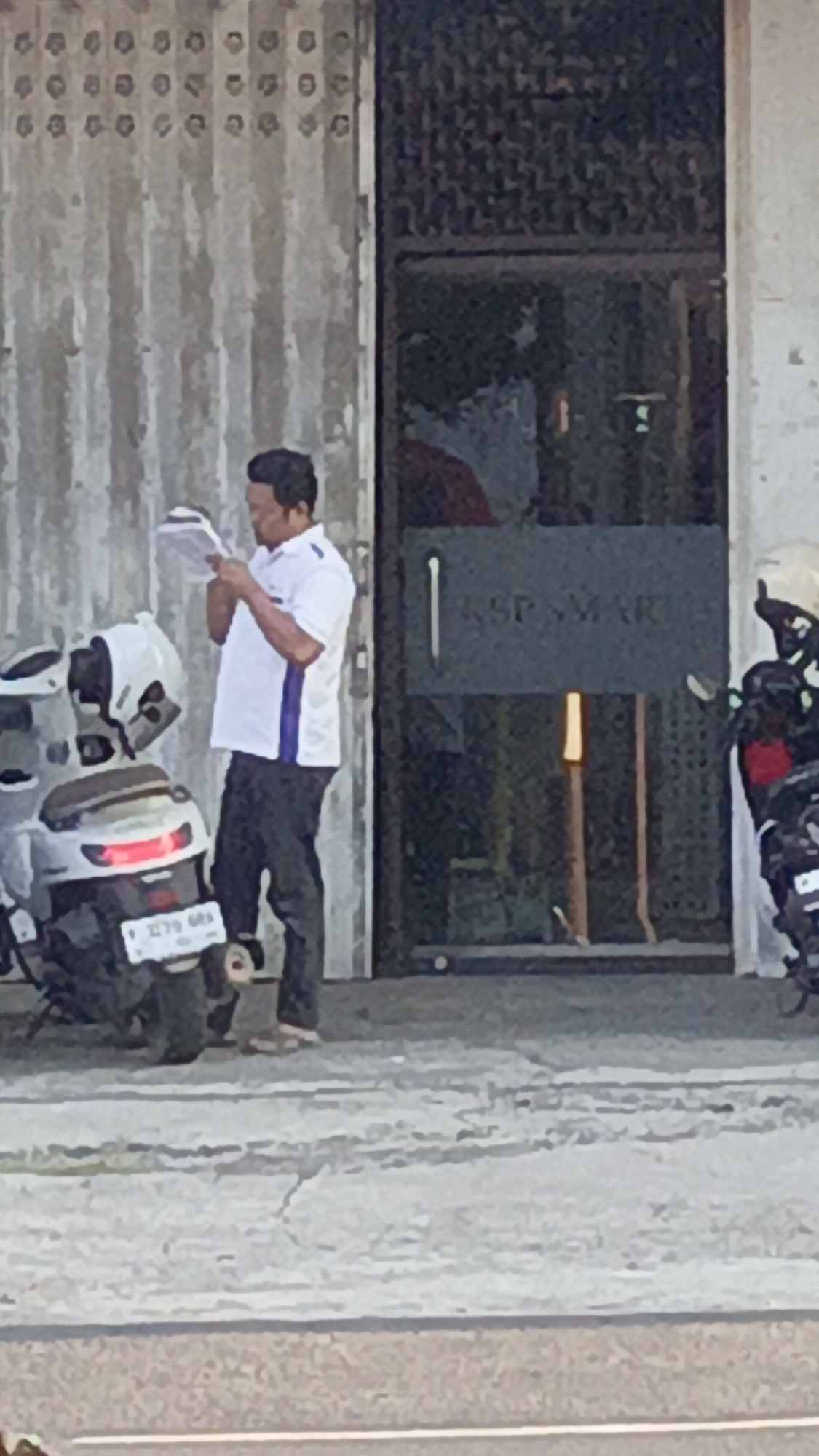 A man wearing a white polo shirt with blue stripes and dark pants stands next to a white scooter in front of a building. He is holding a document and appears to be reading it. The building has a corrugated metal facade and a glass door with text on it. Another motorcycle, partially obscured, is parked to the right of the building. The scene appears to be an outdoor area on a paved surface.