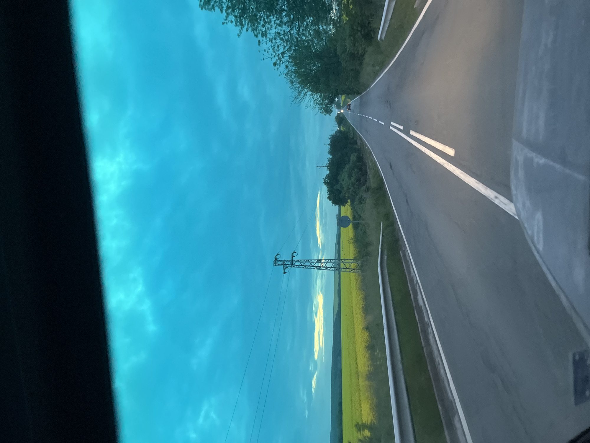 The image shows a view from inside a car driving on a road in Bratovo, Bulgaria. The road is asphalt with white lane markings and is bordered by green fields and trees. An electrical transmission tower stands in the field beside the road. The sky is blue with some clouds. The image is tilted and includes part of the car's interior in the foreground. Another vehicle is visible far ahead in the distance.