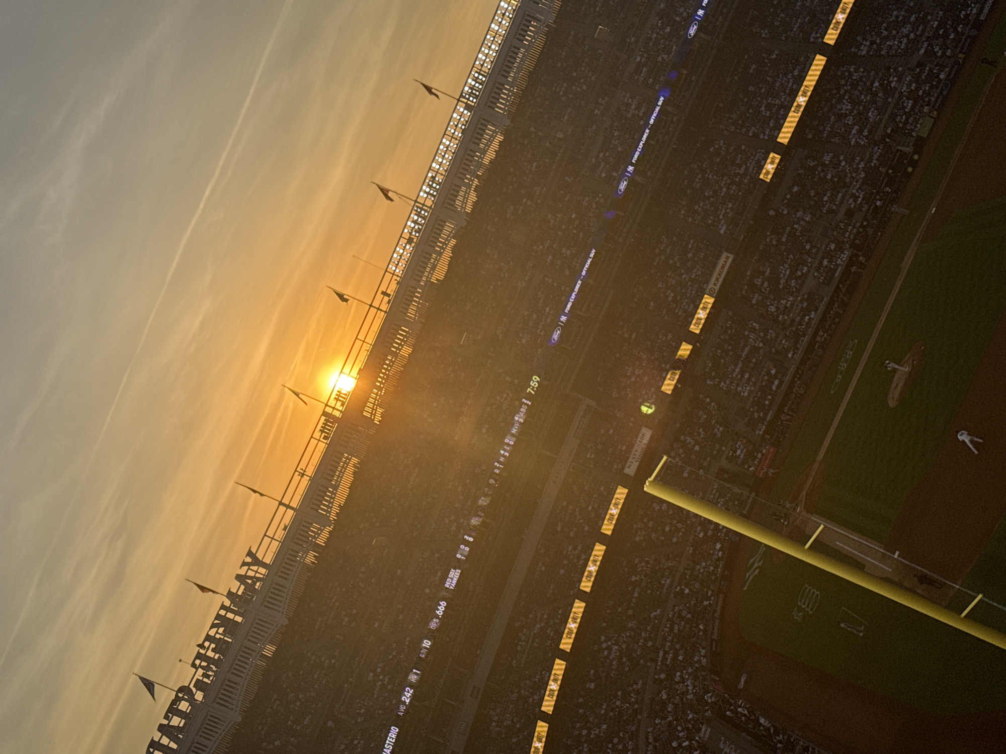This image depicts Yankee Stadium in the Bronx, New York City, United States of America, during sunset. The perspective is from an elevated position, looking down and across the baseball field at an oblique angle. The setting sun is a bright, intense orange orb positioned just above the upper deck of the stadium on the right, casting a warm, golden light and strong glare across the scene. The sky transitions from vibrant orange and yellow at the horizon to a lighter blue-grey higher up, featuring faint contrails.
In the upper left, the white upper deck structure of Yankee Stadium is visible, with the words "YANKEE STADIUM" clearly legible in large letters. Several flags fly from poles along this structure. Stadium lights are illuminated along the upper deck, appearing as a row of bright dots.
Below the upper deck, a large horizontal LED scoreboard displays game information. Visible text includes player statistics such as "MASTERIO AVG .242 HR 1 RBI 10 OPS .666". Below this, team scores are shown as "RED SOX 0" and "YANKEES 0". The time "7:59" is also displayed. Other digital banners feature sponsor logos like "FORD EXPLORER - OFFICIAL SUV", "COOK UNITY", "KIKKOMAN", and "STATE SIDE VODKA".
The baseball field occupies the lower portion of the image. A prominent yellow foul pole extends diagonally from the lower left towards the center. On the field, a pitcher is visible on the pitcher's mound, and another player is near what appears to be first base. A third player is visible near home plate. The field exhibits distinct green grass and brown dirt areas.
The stadium stands are densely populated with spectators, visible as a mosaic of individuals throughout the seating sections, particularly in areas less affected by the direct sun glare. The overall environment is one of an active, populated sporting event illuminated by the dramatic light of a sunset.