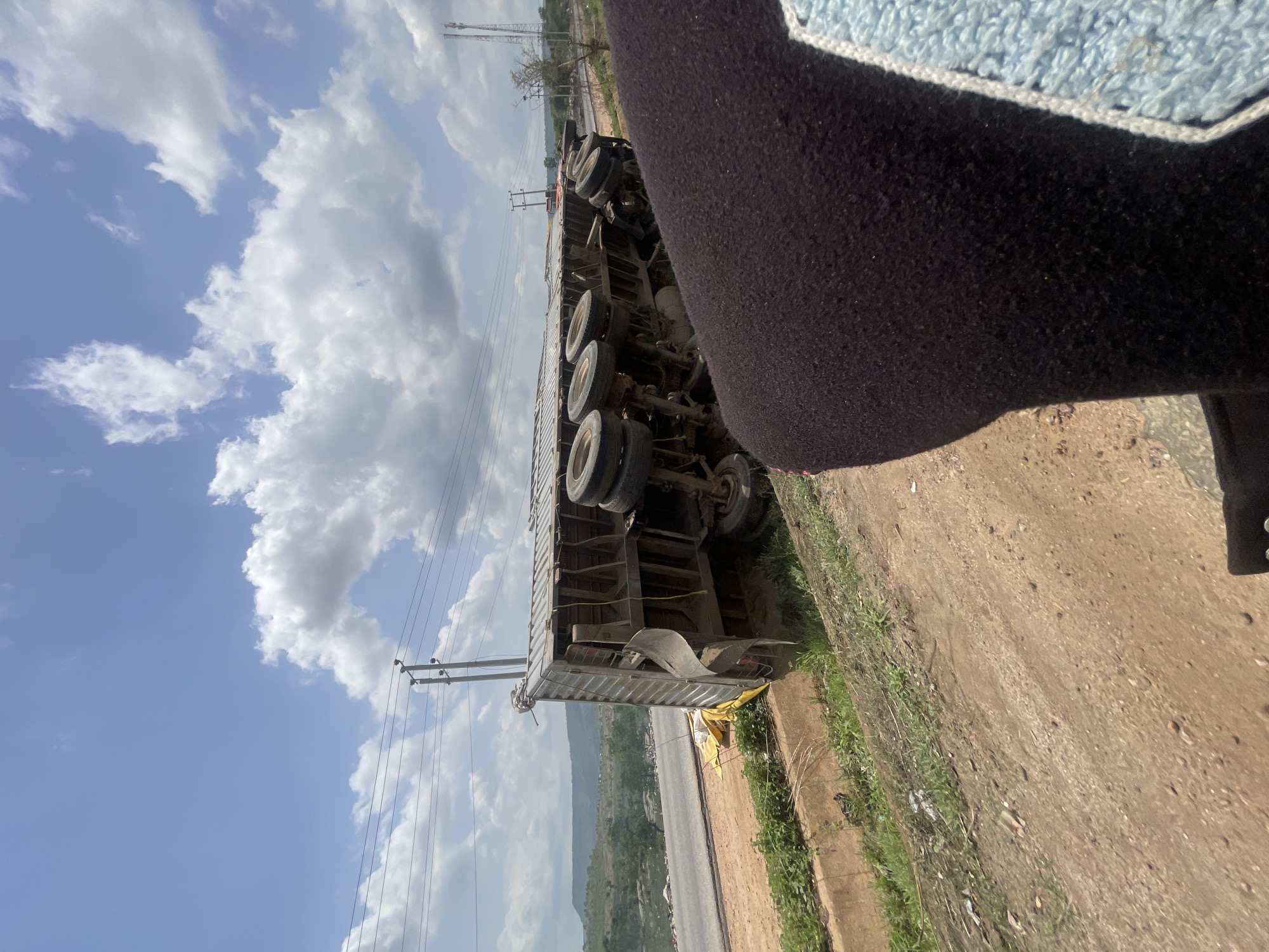 The image shows an overturned articulated truck on a dirt shoulder of a road. The truck is silver and appears to be a cargo carrier. The truck's undercarriage and multiple wheels are visible. Power lines run parallel to the road. A section of highway with a distant view of mountains and greenery can be seen behind the truck. The sky is blue with white clouds. Part of an individual's clothing, possibly a dark jacket or sweater, is visible in the foreground. The image does not specify any interactions or people nearby. The location is suggested to be Lokoja, Nigeria, but that is not evident from the image itself.
