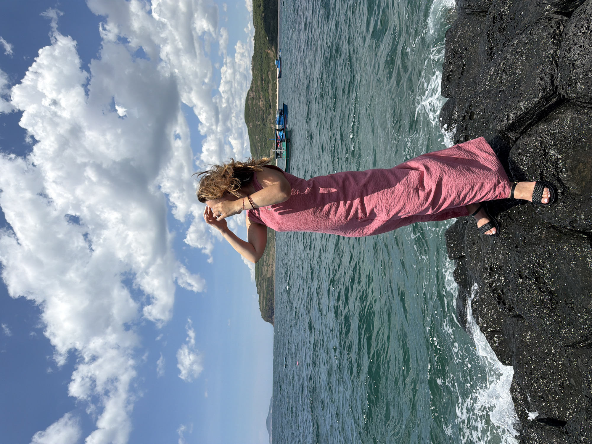 A woman with windswept brown hair stands on dark, wet rocks by the sea. She is wearing a textured, rose-pink, sleeveless dress and black sandals. Her hands are raised to her head, as if shielding her eyes from the sun or wind. The ocean is a deep blue-green, with white waves crashing against the rocks. In the distance, there are several small boats on the water and a green, hilly coastline with buildings visible. The sky is bright blue with scattered white clouds. The image appears to be taken on a sunny day. The specific location is the shore of Phú Lương City, Vietnam.