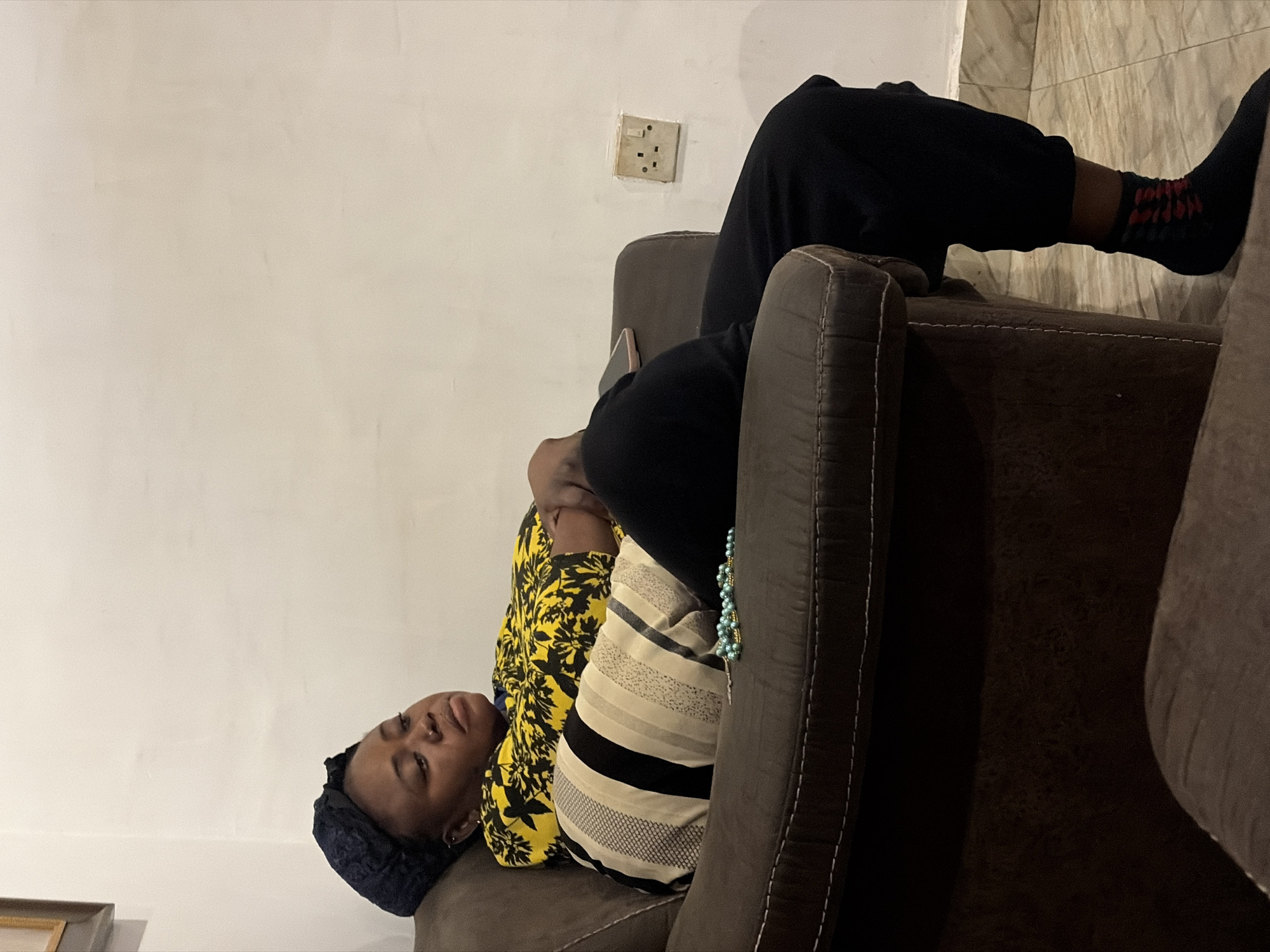 A woman is sitting on a brown armchair in a room. She is wearing a yellow and black patterned top, black trousers, and black socks with red accents. She has a dark blue headwrap on. Her arms are crossed in front of her, and her eyes are closed. A string of light blue beads is on the armrest of the chair. The room has a white wall with a power outlet and a light switch. The floor is tiled. The image appears to be taken indoors in a residential setting within the city of Jos, Nigeria.