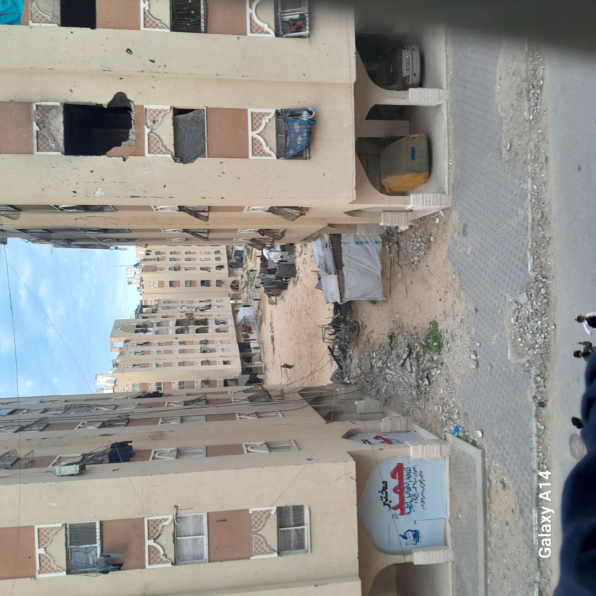 The image portrays a heavily damaged urban residential complex in Khan Yunis, Palestinian Territory, viewed from an elevated perspective. Multi-story, light-colored apartment buildings with brown and white decorative elements flank a central, unpaved open area.
The buildings exhibit extensive damage, including numerous impact craters and bullet holes on their facades. Many windows are shattered or completely blown out, revealing dark interior spaces, while others are covered with dark tarps or hanging fabric. Some windows retain their grates, partially obscured by debris. On the ground level of the left building, a large banner in Arabic advertises "Hamad Laboratory" (مختبر حمد) and "for all medical tests" (ل كافة التحاليل الطبية). The right building features arched ground-level openings, under which two cars are parked: one silver and uncovered, the other covered with a tan tarp.
The central area between the buildings is an unpaved expanse of sand and dirt, littered with concrete rubble, twisted metal, and miscellaneous debris. Several makeshift shelters, constructed from tarps and salvaged materials, including one with a visible UNICEF logo, are erected across this central ground.
Multiple small figures are visible within the scene. One individual walks across the sandy area towards the left, while a small group stands near the distant shelters. In the immediate foreground, two partially obscured individuals are seen on the paved street. The sky above is partly cloudy, and numerous black electrical wires traverse the scene between the buildings.