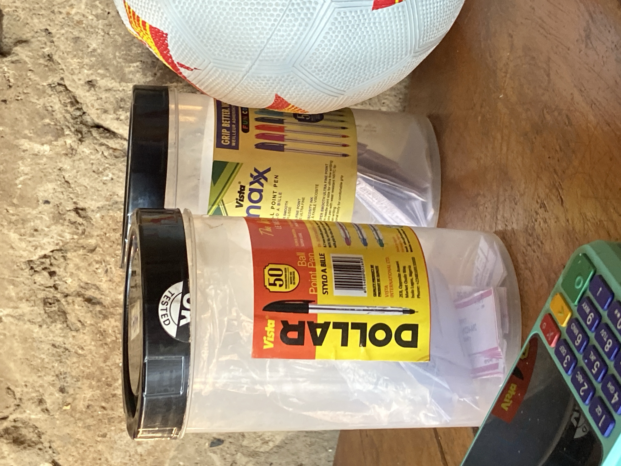 In Mararaba, Nigeria, on a wooden surface, a close-up shot captures a still life arrangement. Two clear plastic containers, each with a black lid, are prominent. The container on the left displays a yellow label with red text that reads "DOLLAR" vertically, and a smaller label indicating "50 Ball Point Pen" with an image of a pen. Inside this container, white slips of paper are visible. The second container, to the right, features a yellow label with text including "Vista max" and "GRIP BETTER. W...". Several colored pens are visible within this container. To the right of the containers, a portion of a white and red soccer ball is visible. In the foreground, a corner of a mint green electronic device with a purple keypad is visible. The background consists of a rough, textured stone wall.
