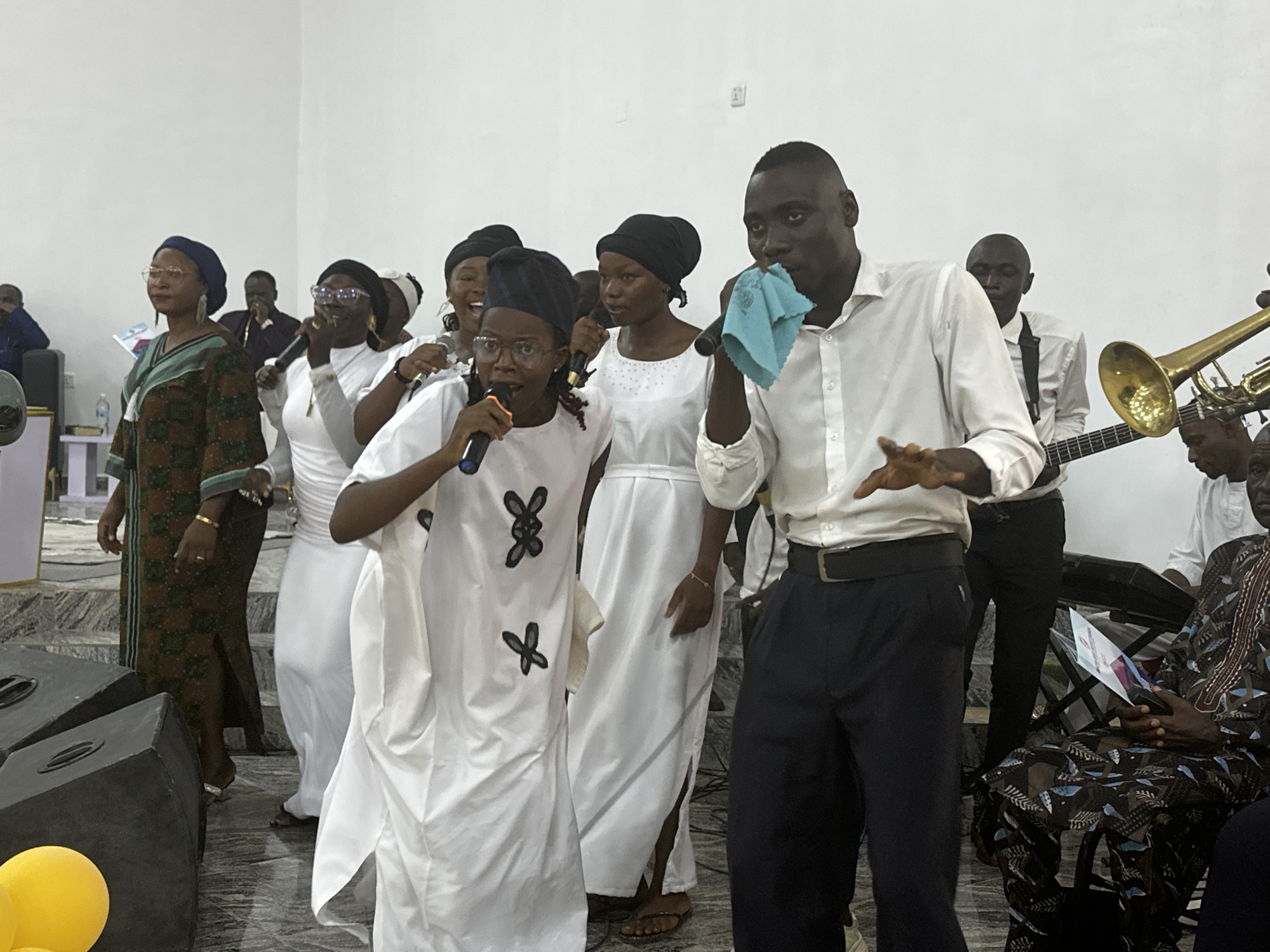A group of people are performing, likely in a church or auditorium. The location is likely Goni Gara, Nigeria. Several singers are in the foreground, wearing white robes or shirts. One man is holding a blue cloth. Behind them are musicians with instruments, including a trombone and a bass guitar. Audience members are seated on the right and further in the background. The setting appears to be indoors with white walls and a tiled floor. There are speakers on the left side of the image. The lighting seems to be diffused and somewhat bright.