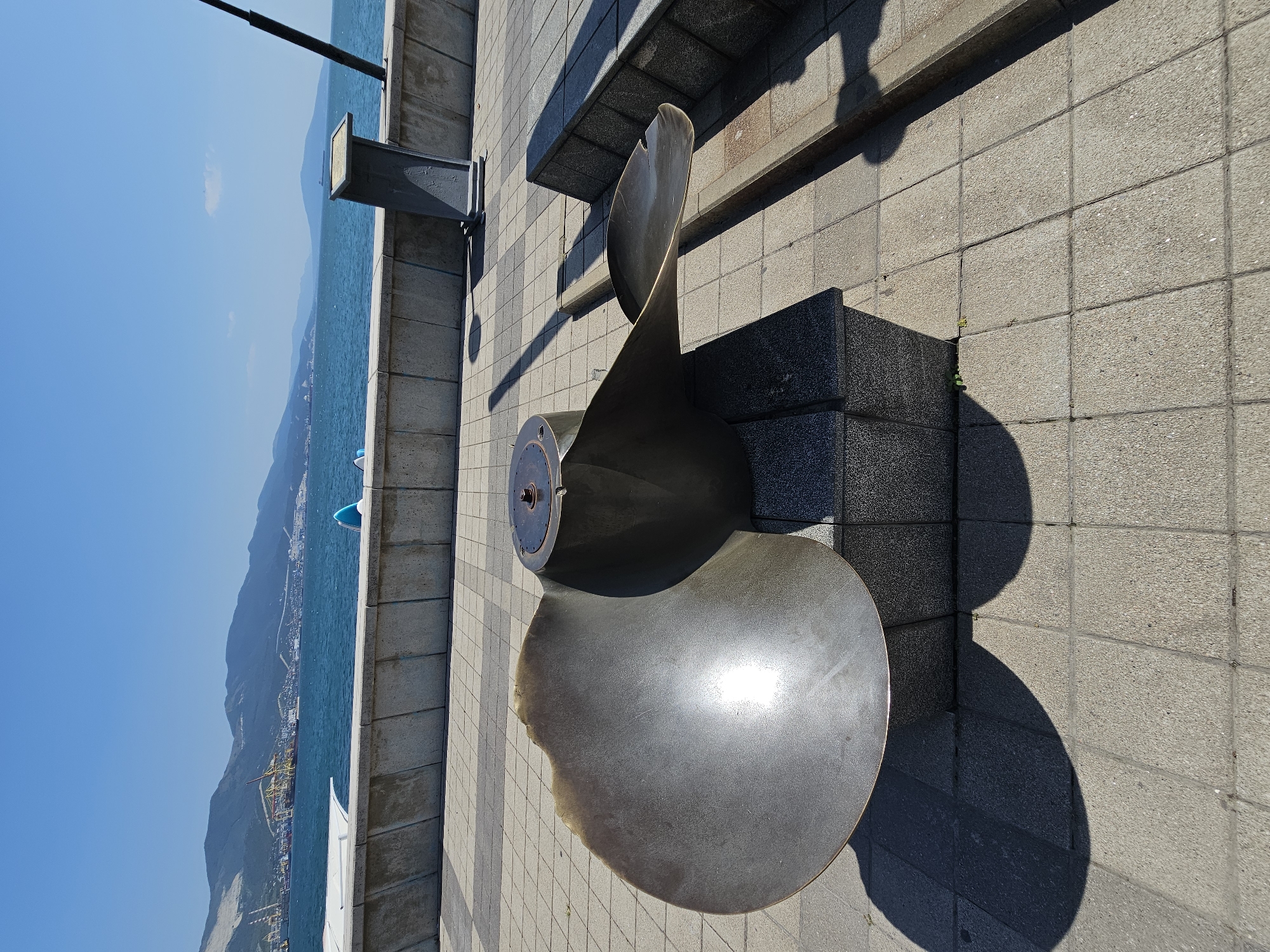 This is a wide, eye-level, outdoor, daylight photograph of a large metal ship propeller monument situated on a paved promenade overlooking the sea in Novorossiysk, Russia. The propeller is dark bronze colored and weathered, with one blade prominently displayed, curved and facing left. It rests on a stepped granite pedestal. Behind the monument, a concrete seawall runs horizontally across the frame, with a metal informational display stand visible to the right of the propeller. Beyond the seawall, the turquoise sea stretches towards a distant coastline lined with mountains and industrial port structures, including cranes. The sky is a clear, bright blue. The paved promenade is made of light-colored square tiles, with shadows from the propeller and other unseen objects casting distinct shapes on the ground. The environment suggests a public area, likely a tourist attraction or memorial, in a coastal city.