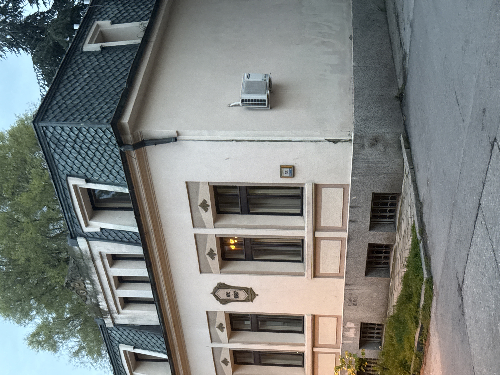 This media file captures an exterior view of a multi-story building, likely a house or a small residential block, situated along a paved street in Varna, Bulgaria. The building features a cream-colored facade with decorative elements around its windows, and a distinctive dark gray or black tiled roof with dormer windows.
Several windows are visible, including those on the upper floors and a series of basement windows fitted with protective metal grates. An outdoor air conditioning unit is mounted on the upper wall of the building. Green trees and some grass line the side of the street next to the building.
Visible text includes a small rectangular plaque displaying the number "33" near an upper window, and a more ornate, shield-shaped plaque on the main facade that reads "GTC 1887". A small digit "0" is also visible on the display of the air conditioning unit.
The scene appears to be set during daytime, possibly late afternoon or early evening, as an interior light is illuminated in one of the windows, casting a warm glow. There are no people visible in the image. The sky is bright but appears overcast or partly cloudy.