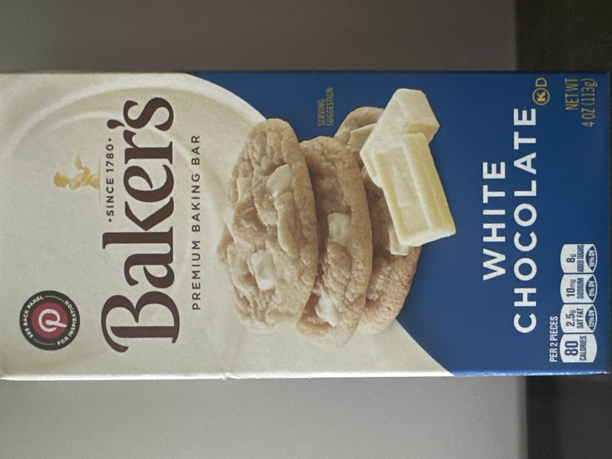 The image shows the front of a Baker's White Chocolate Baking Bar box. The box is primarily white with blue accents at the bottom. The brand name "Baker's" is prominently displayed in large, dark brown lettering. Below the brand name, it reads "PREMIUM BAKING BAR." In the center of the box, there are images of white chocolate chip cookies and white chocolate chunks. Nutritional information is visible at the bottom left, indicating "PER 2 PIECES" with values for calories, fat, sodium, and added sugars. The box also displays the net weight as "4 OZ (113g)" and a kosher certification symbol. The image is taken from a slightly angled, eye-level perspective, with a plain, neutral background. The location of the image within Lower Paxton Township, United States of America, cannot be determined from the visual information provided in the image itself.