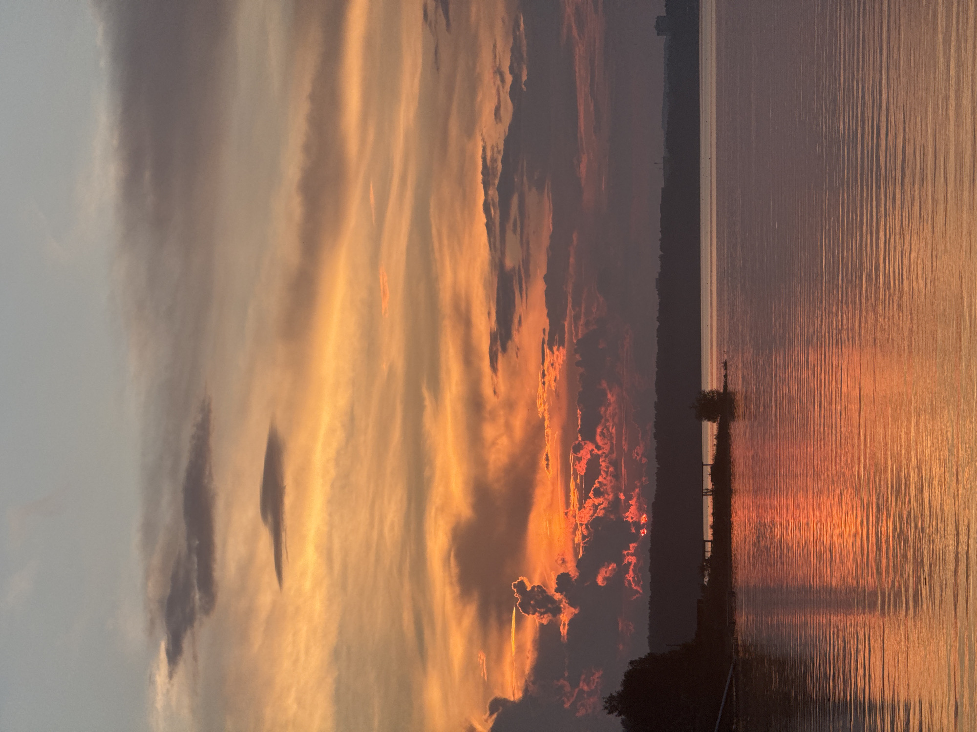 The image depicts a tranquil sunset over the Kama River in Chaikovsky, Russia. The sky is a vibrant canvas of oranges, reds, and purples, with wispy clouds catching the last rays of the sun. The water reflects the colorful sky, creating a shimmering, ethereal effect.
On the left bank of the river, a dense forest line is silhouetted against the bright sky. A small structure, possibly a pier or a boathouse, is visible on the water's edge, adding a touch of human presence to the otherwise natural landscape. The overall atmosphere is one of peace and serenity, with the gentle ripples on the water and the warm hues of the sunset creating a soothing and picturesque scene. The distant horizon line suggests a vast expanse of water, disappearing into the hazy glow of the setting sun.