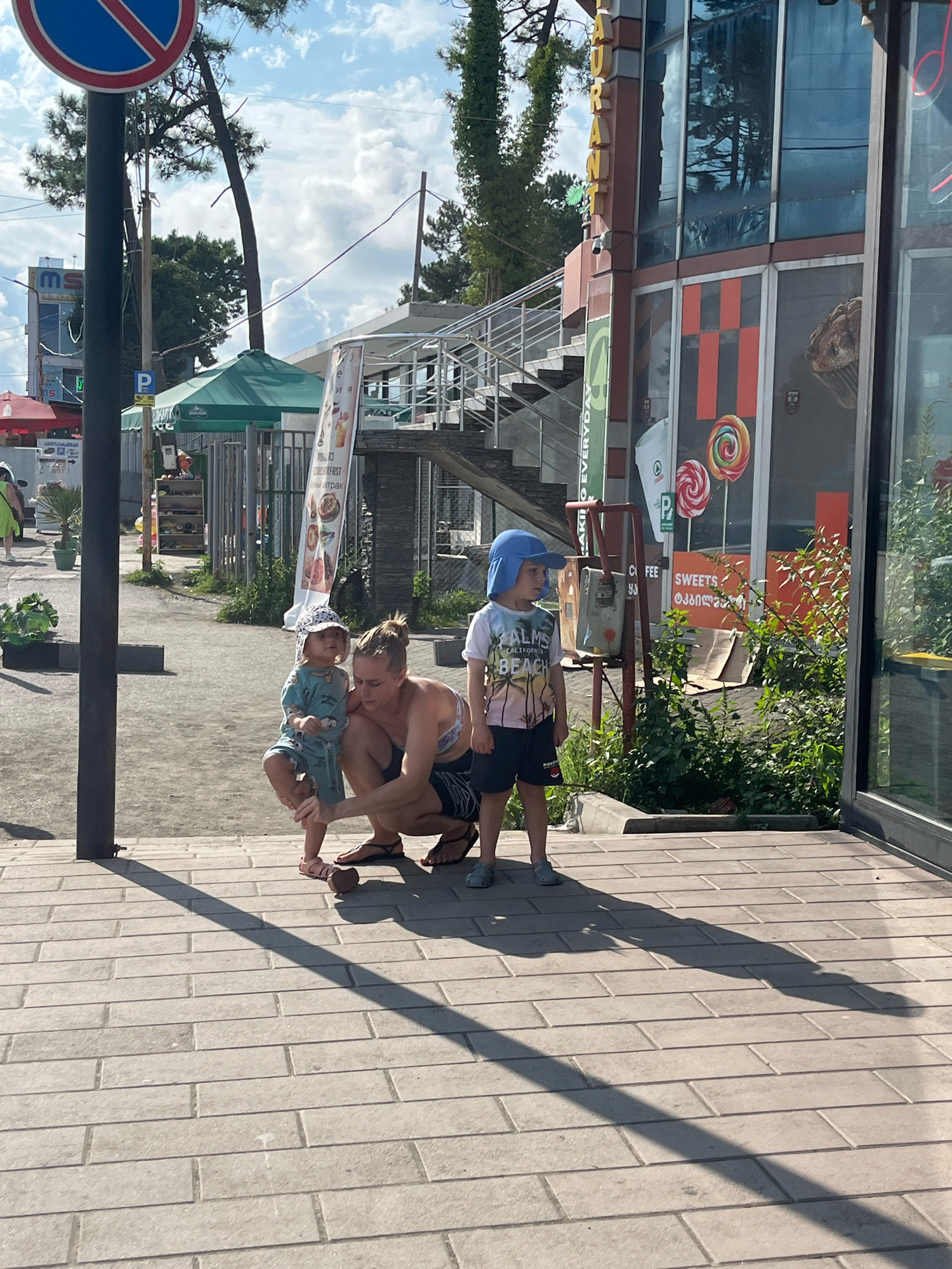 In Ureki, Georgia, on a sunny day, a woman kneels on a paved walkway in front of a building with large glass windows. She is wearing a dark tank top and dark shorts. To her left, a young child in a blue patterned outfit and a sun hat stands, and the woman appears to be adjusting the child's shoe. To the woman's right, another young child wearing a blue hat, a t-shirt with a beach scene, and dark shorts stands and looks to the left. The building behind them has a sign with colorful lollipops, and the words "SWEETS" are visible. In the background, there is a staircase leading up to a walkway, and some trees and a blue sky with clouds. A street sign with a red circle and diagonal line is visible on a pole in the foreground.