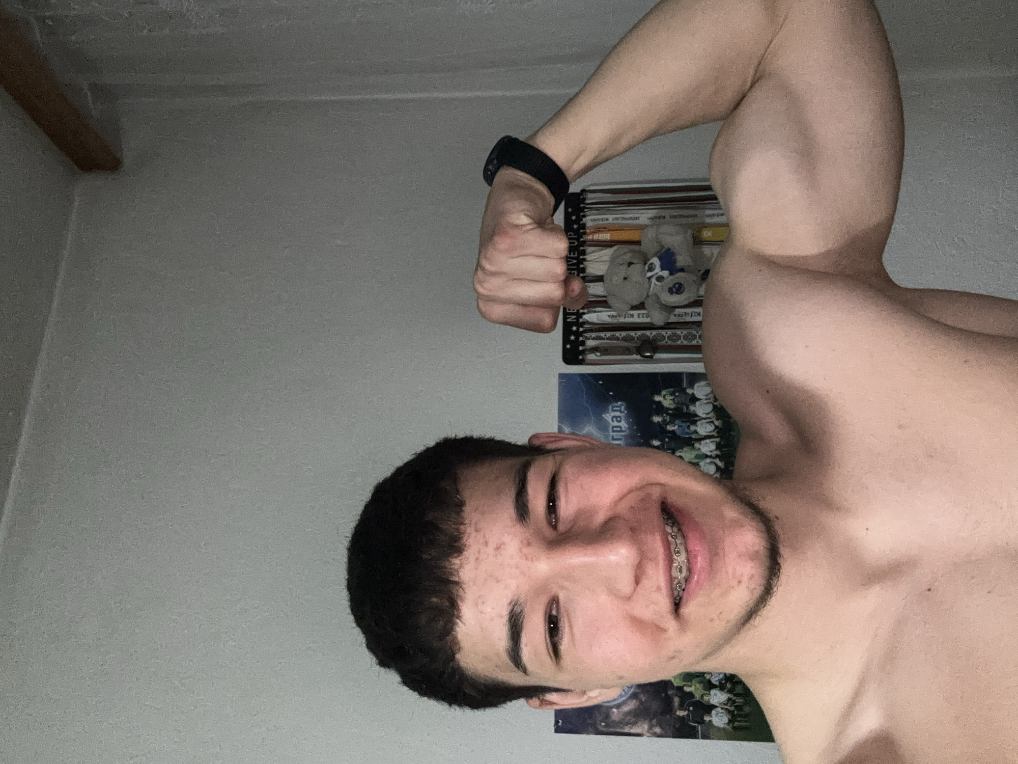 A shirtless young man with braces is posing and flexing his right arm. He is smiling. He wears a black fitness tracker on his right wrist. Behind him is a shelf with items on it including a teddy bear. Below the shelf is a poster with the word "град" on it. The image was likely taken indoors. The location is impossible to determine, due to the angle of the photo and lack of external information.