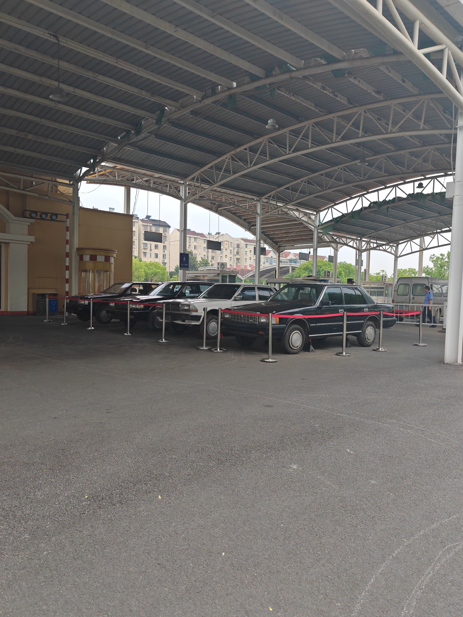 The photograph shows a row of four vintage black and white cars lined up under a large metal structure resembling a carport or open-air pavilion. The vehicles are situated on an asphalt surface, separated from a building on the left and possibly from other vehicles on the right by a red velvet rope barrier system with silver poles.
In the background, partially visible, there are residential buildings with multiple stories. A figure can be seen standing near a van or larger vehicle further back. The architecture of the structure overhead includes exposed metal beams and a corrugated roof.
The image suggests a display or exhibition of vintage automobiles in an outdoor or semi-enclosed setting. The location is the Xuanwu District in China.