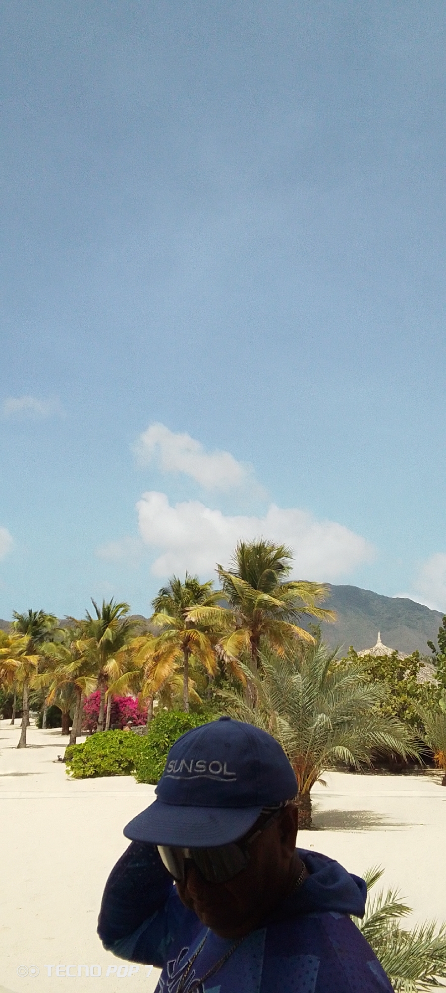 A sunny day in Pedro González, Venezuela. Palm trees and other tropical vegetation line a light sandy area. In the foreground is a person wearing a dark blue baseball cap with "SUNSOL" printed on it and sunglasses, with a blue patterned shirt and a gold chain. In the background, a hill is visible in the distance. There are scattered white clouds in the blue sky.