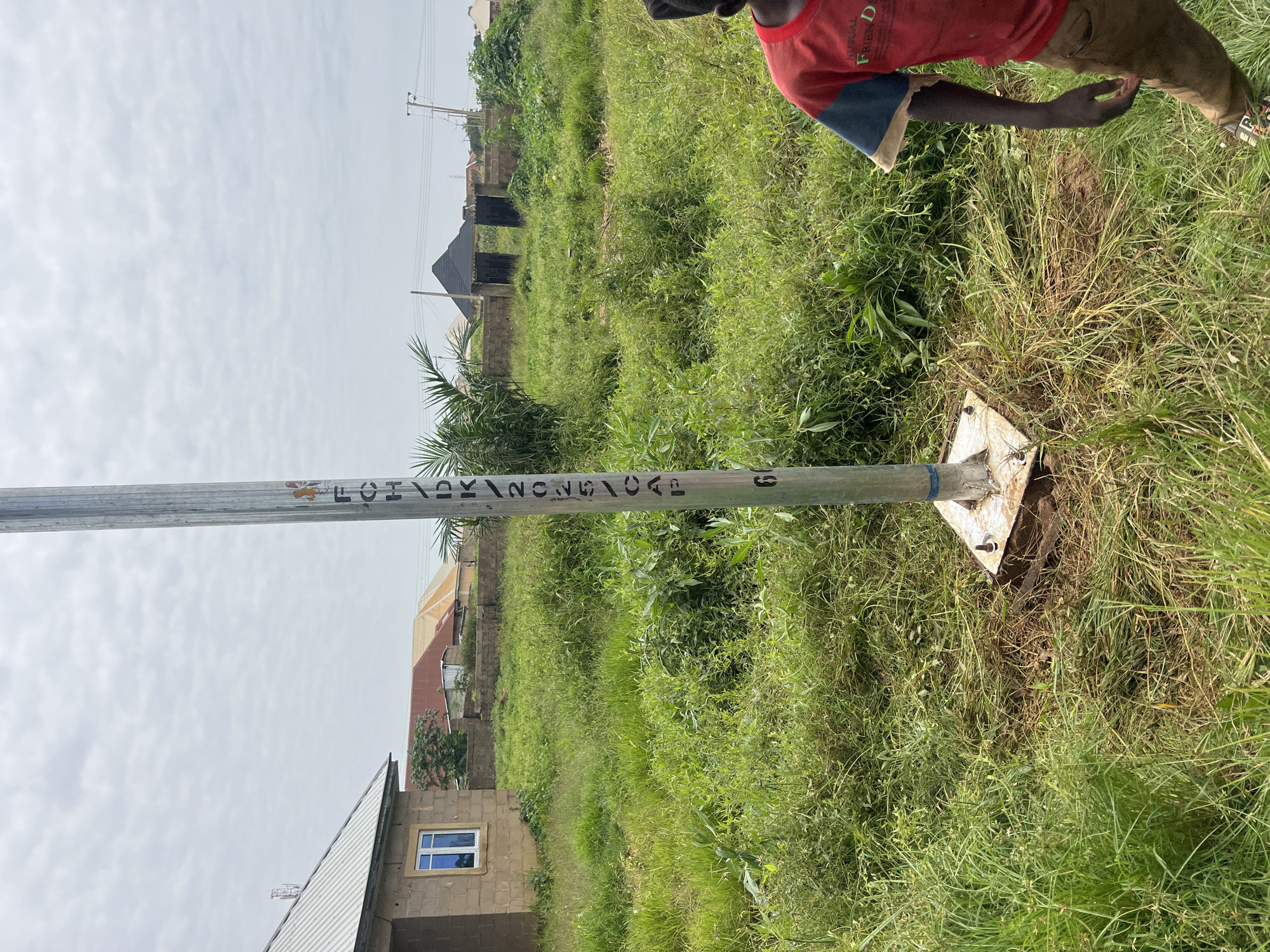 The image depicts an outdoor scene in a grassy, undeveloped area that is likely within the city of Jos, Nigeria. A tall, thin metal pole stands prominently in the foreground, anchored to the ground by a square metal base with bolts. Markings, possibly alphanumeric characters and a number "6", are visible vertically along the pole. To the left of the pole is a portion of a building constructed with light brown bricks, featuring a grey, corrugated metal roof and a single window with blue panes. Further in the background, several other structures with various colored roofs are visible, along with trees and vegetation. The sky is overcast with a cloudy, pale gray sky. In the right foreground, a person, a young boy, is partially visible from the chest down, wearing a red t-shirt with blue sleeves and brown trousers. He is standing in the grass, facing away from the camera and towards the right. The overall impression is of a semi-urban or developing area with natural elements and some signs of construction.