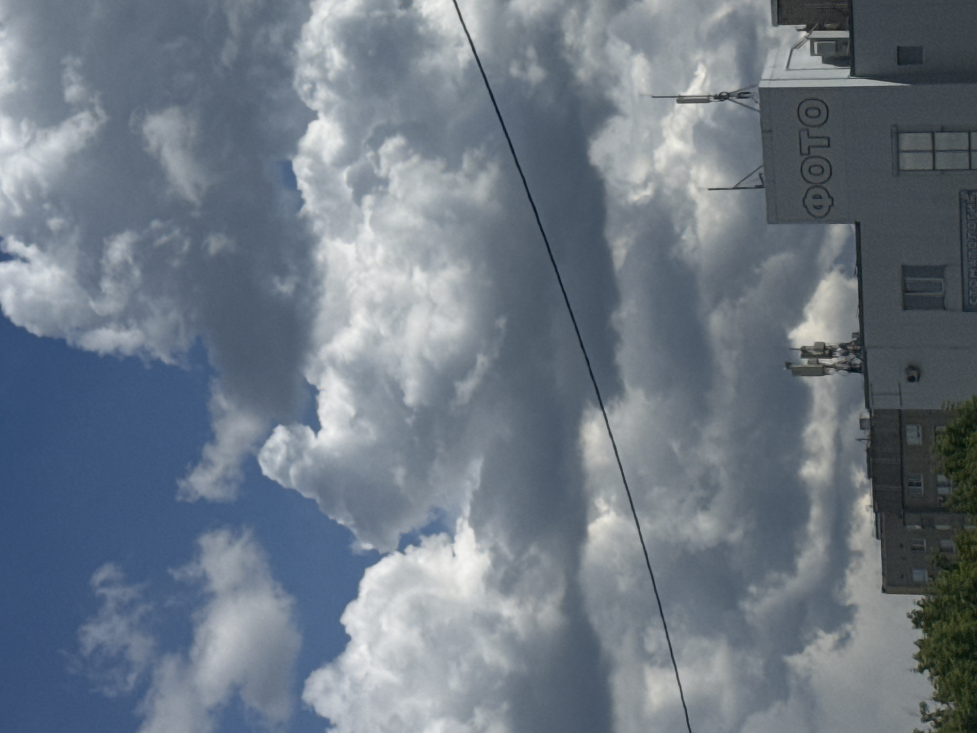 The image depicts a building with a sign reading "ФОТО" (photo in Russian) on its side, interspersed with clouds in the sky. A power line runs across the image. The location is in an urban area. The building appears to be residential, with multiple windows. Trees are visible on the right side of the image. The sky is a mixture of blue and cloudy.