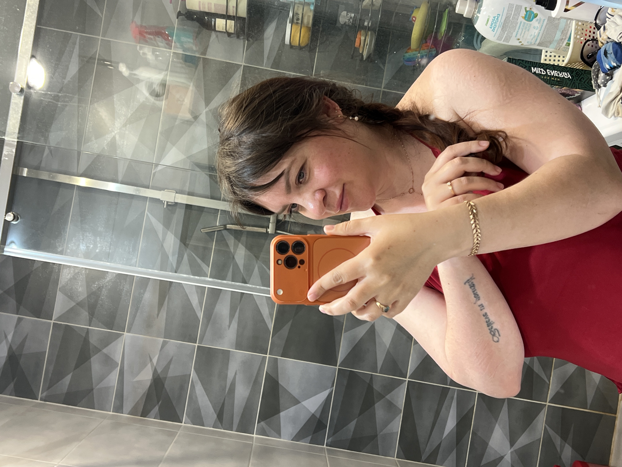 A woman with brown hair in a braid takes a mirror selfie in a bathroom. She is wearing a red top, a gold necklace, a gold bracelet, and a ring on her left hand. Her right arm, which is holding an orange phone, has a tattoo. The bathroom has gray tiled walls and a glass shower door. Shelves in the background contain various bottles and containers. The setting is likely a residential bathroom in Noviy, Russia.