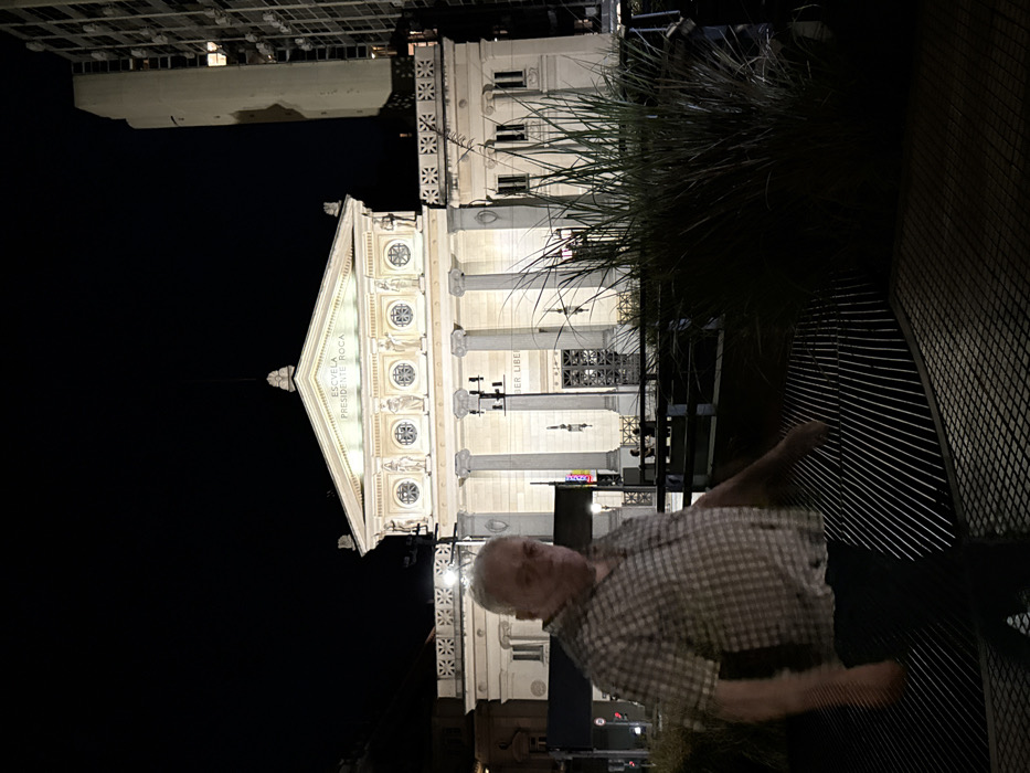 A rotated night shot shows a man in a plaid shirt and dark pants walking across a metal fence or structure. Behind him, the Escuela Presidente Roca building is brightly lit, displaying a classical facade with columns and arched windows. Above it, another building is visible in the darkness. Some shrubbery is on the right side of the frame.