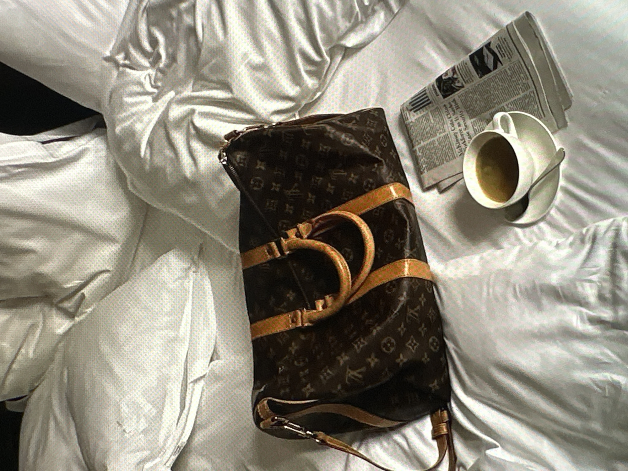 The scene is a high-angle shot of a bed with white bedding. A brown Louis Vuitton duffel bag is placed in the center of the bed. Next to the bag, on the right, is a folded newspaper with a cup of coffee resting on top of it. The presence of these items suggests a state of leisure or recent arrival, possibly in a hotel room. The location in Yerevan, Armenia cannot be determined from the visual information provided.
