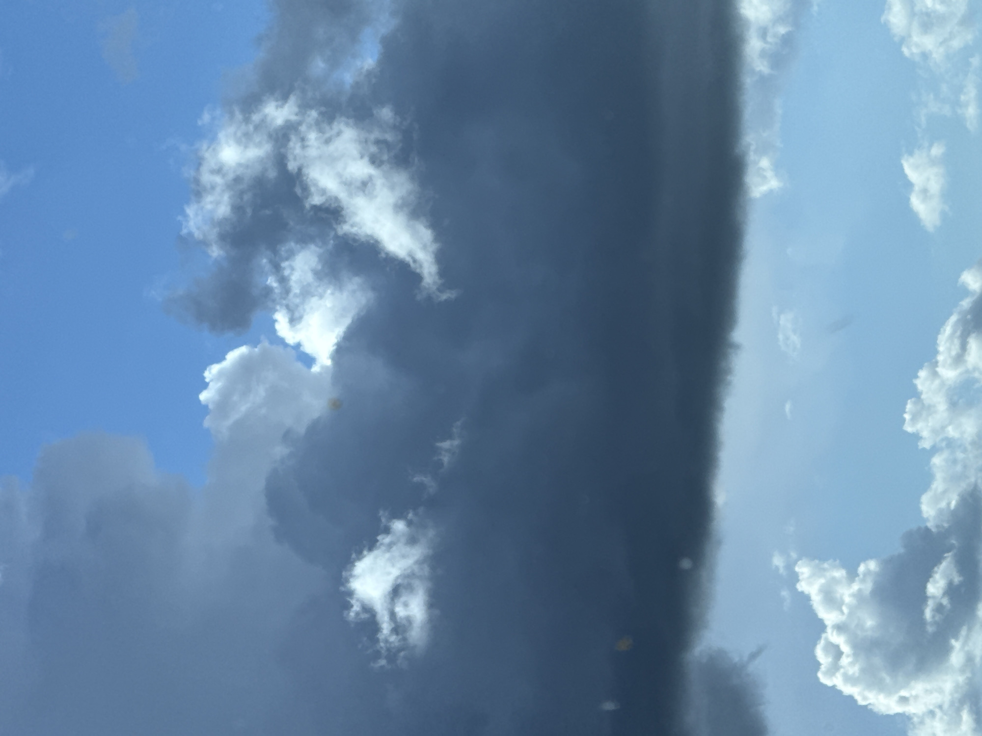 The image depicts a sky with contrasting cloud formations. On the left side, there are cumulus clouds against a blue sky. The clouds are varying in size, density, and color from white to gray. On the right side, a very dark, almost black, vertical band of clouds is observed. Adjacent to this dark band, there are lighter, white clouds against a light blue sky. The image is rotated. The location is Перовское сельское поселение, Ukraine. There are no people or human-related subjects in the frame.