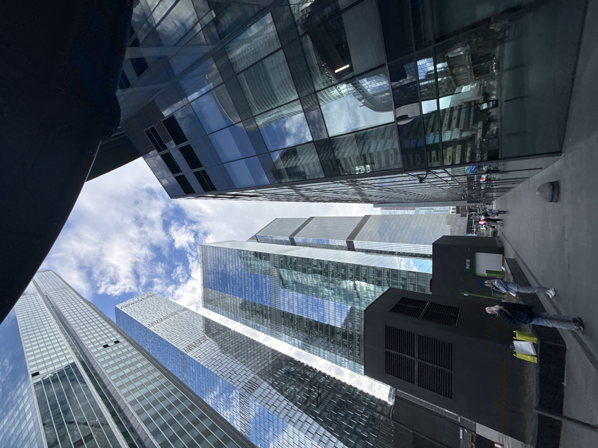 The image depicts a view looking upwards at a cluster of modern skyscrapers in Khimki, Russia. The buildings are characterized by glass facades, reflecting the sky and clouds. A portion of a darker structure or building is visible on the lower left and right side of the view. People are walking on the sidewalk, next to a dark, rectangular, box-like structure, presumably a commercial area, near the base of the buildings. The sky is blue with scattered white clouds.