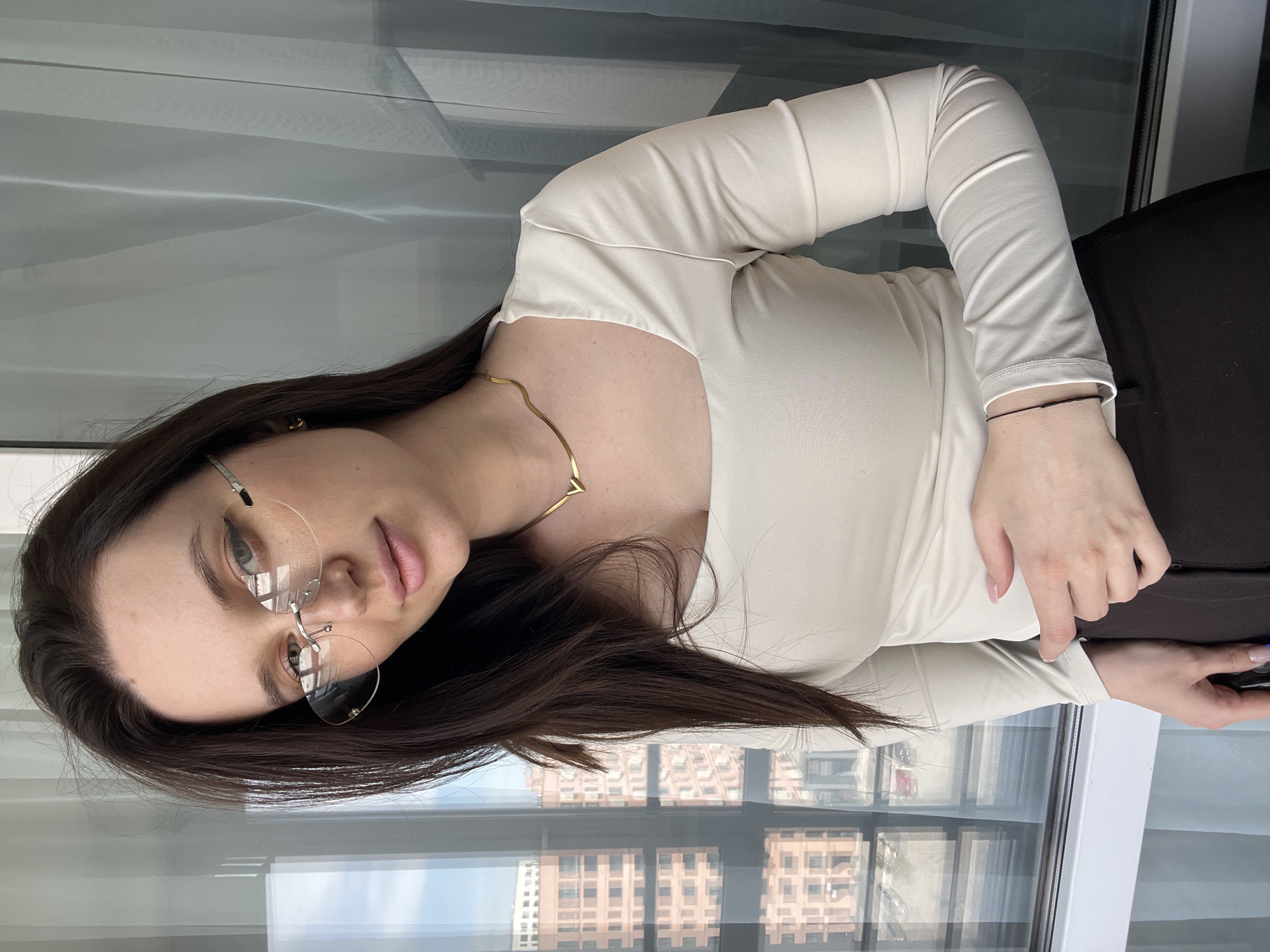 A young woman with long brown hair is seen in a medium shot, wearing rimless eyeglasses, a gold necklace, and a beige, long-sleeved top. She is standing by a window overlooking an urban landscape. The background reveals a cityscape with buildings. Based on the architectural style and the lighting conditions, the location is identified as Tyumen, Russia. The woman is looking towards the camera with a slight smile, her hand resting on a dark brown object.